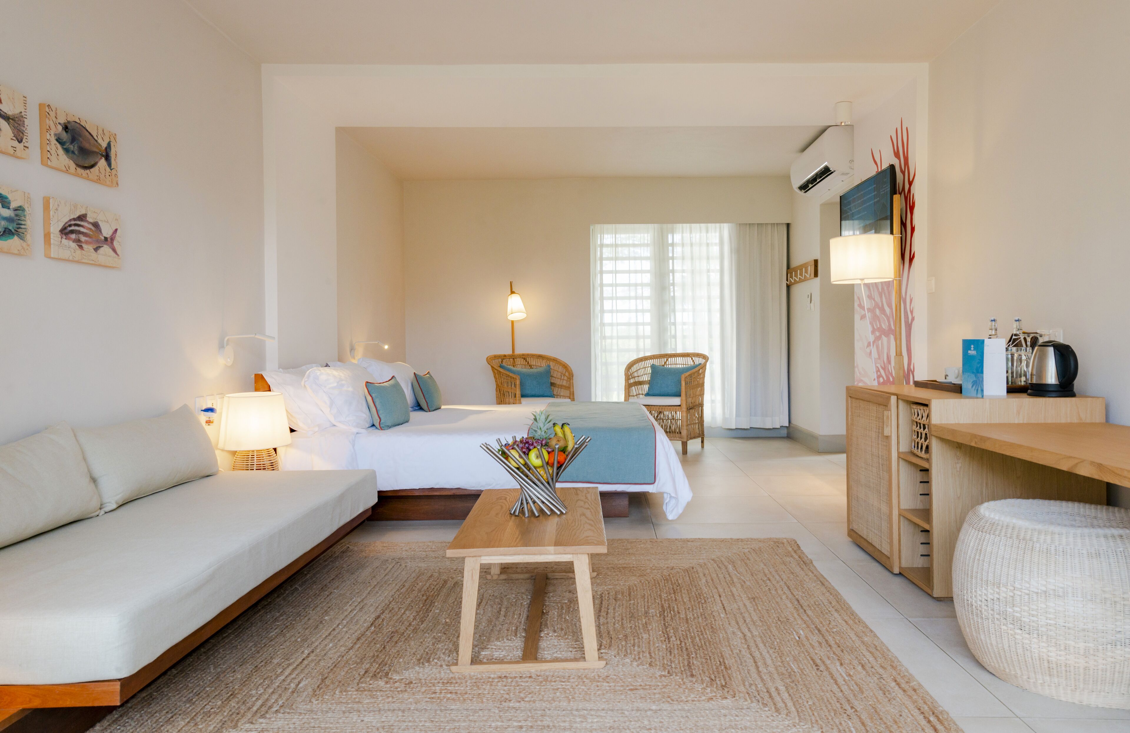 Una habitación de hotel moderna con una cama, sofá, dos sillones de mimbre, una mesa de madera con fruta y una kitchenette.