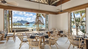 A spacious dining area with wicker furniture and large windows overlooking a beach and ocean.