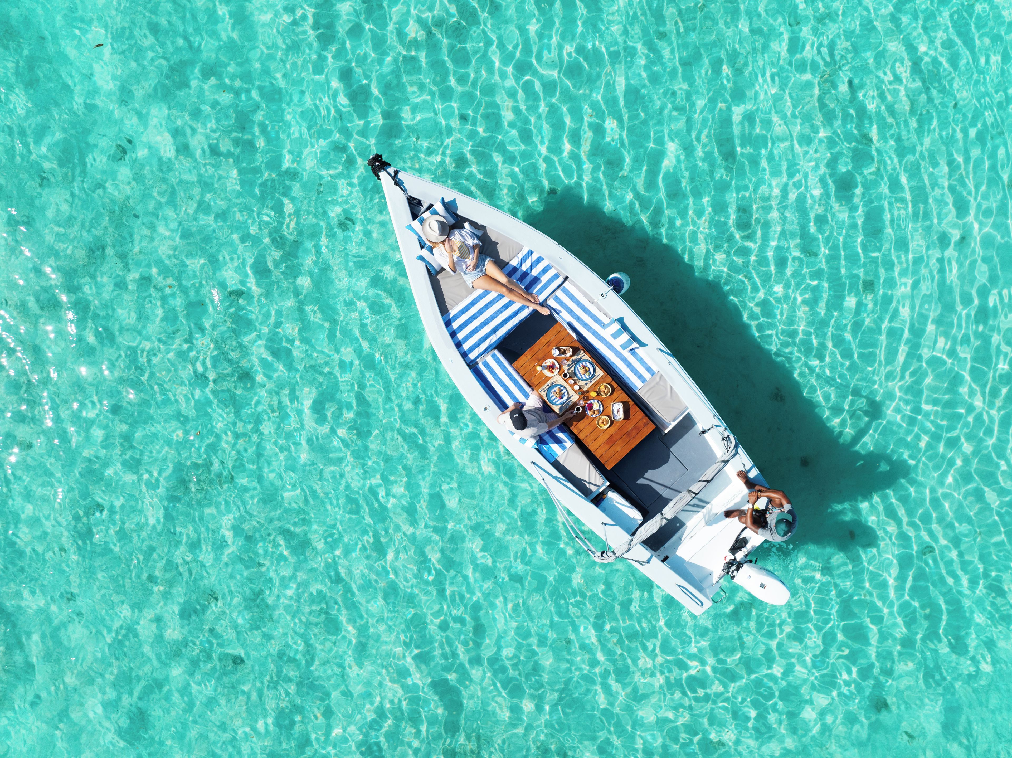 A boat with a dining table and two people seated, floating on clear turquoise water.