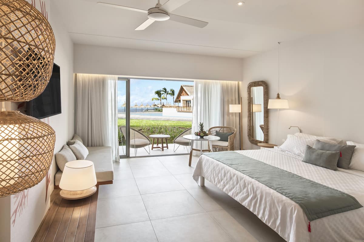 A hotel room with a bed, a sofa, a dining table, and chairs. There is a balcony with a view of the beach and palm trees.