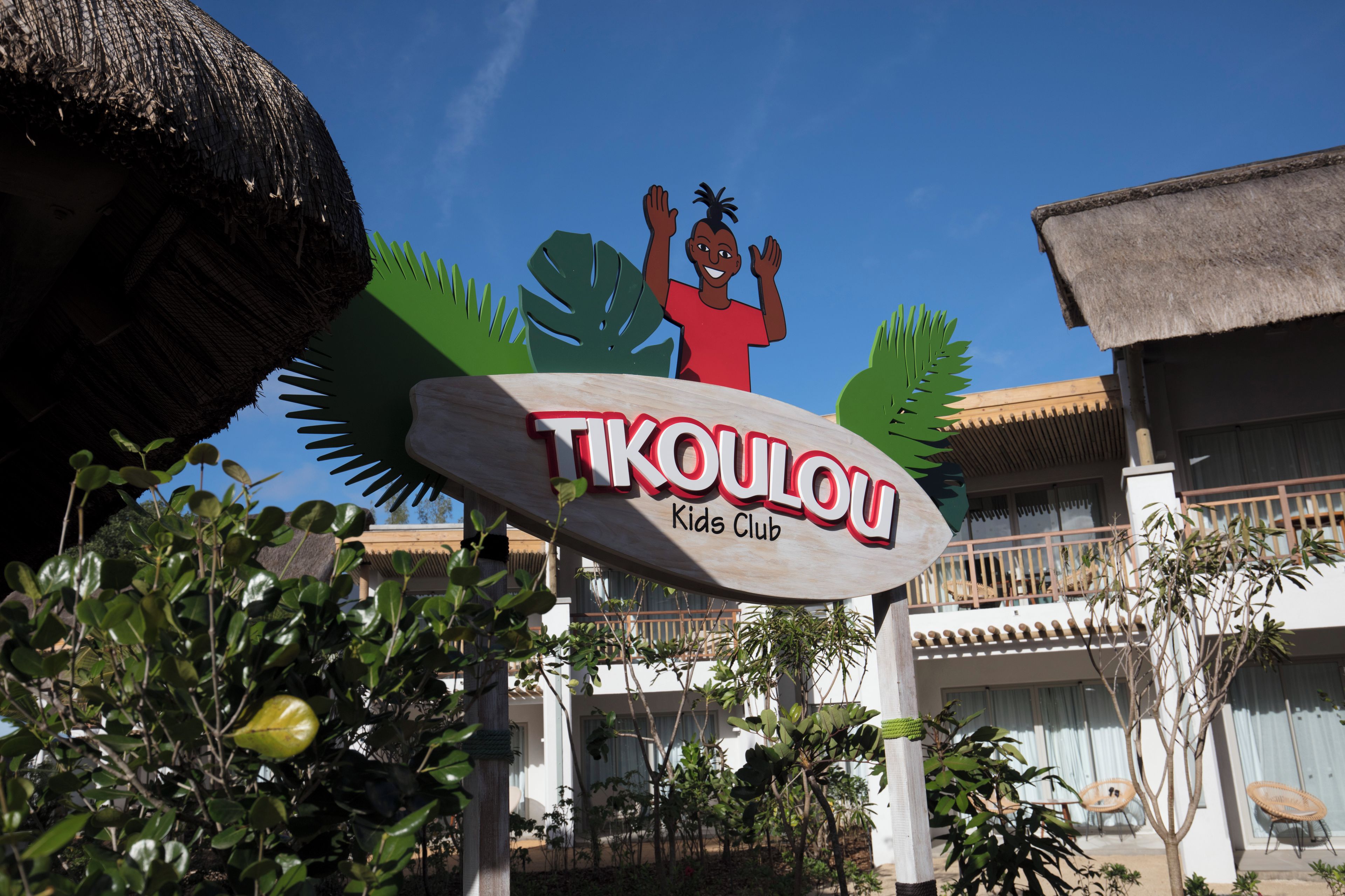 A sign for 'TIKOULOU Kids Club' with a tropical theme, including a stylized palm tree and a child waving.