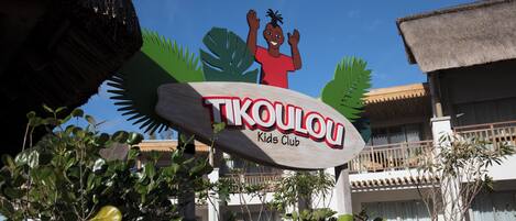 Letrero para el 'TIKOULOU Kids Club' con temática tropical, que incluye una palmera estilizada y un niño saludando.