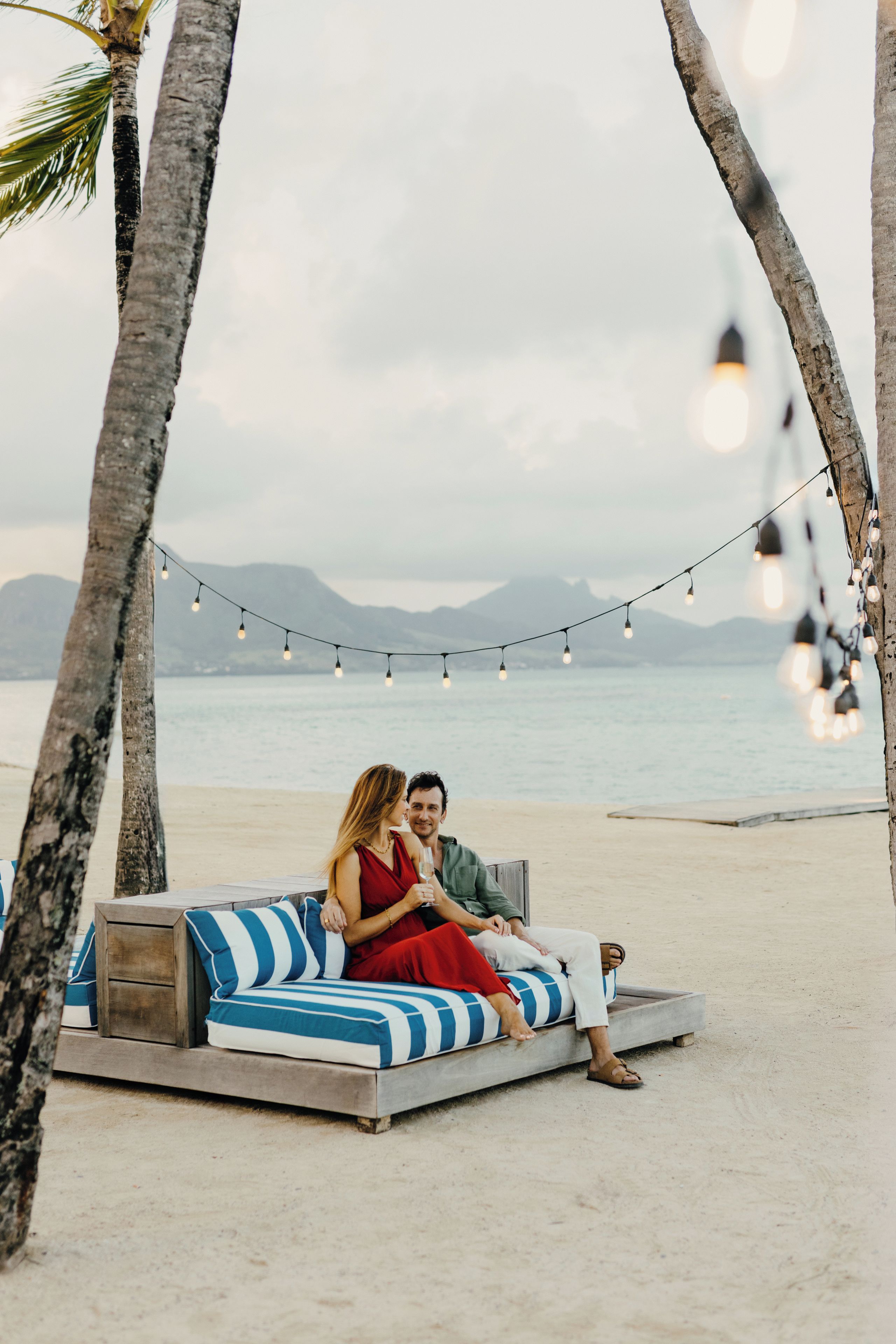 A couple sitting on a beach lounge chair with a view of the sea and mountains in the background.