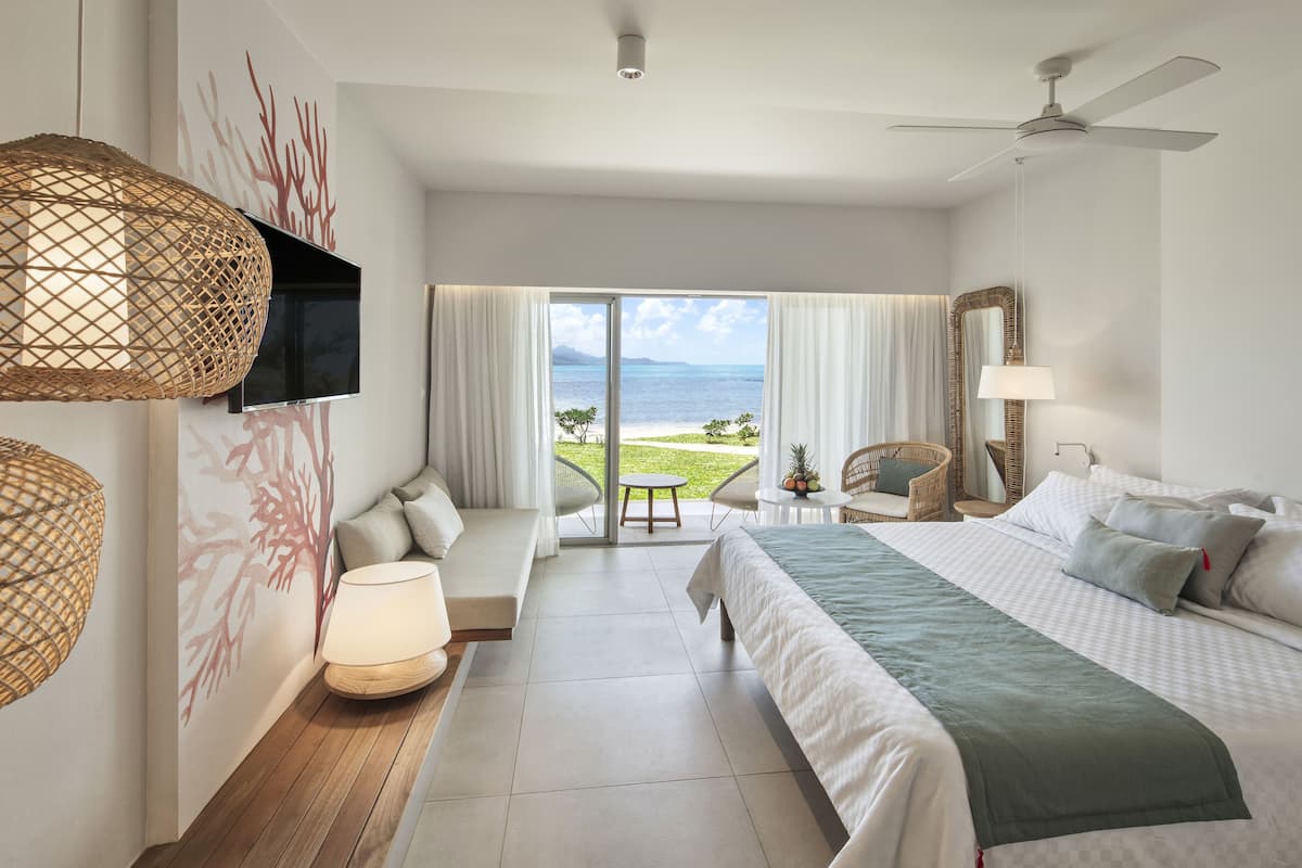 A hotel room with a bed, a sofa, a TV, a ceiling fan, and a view of the ocean.
