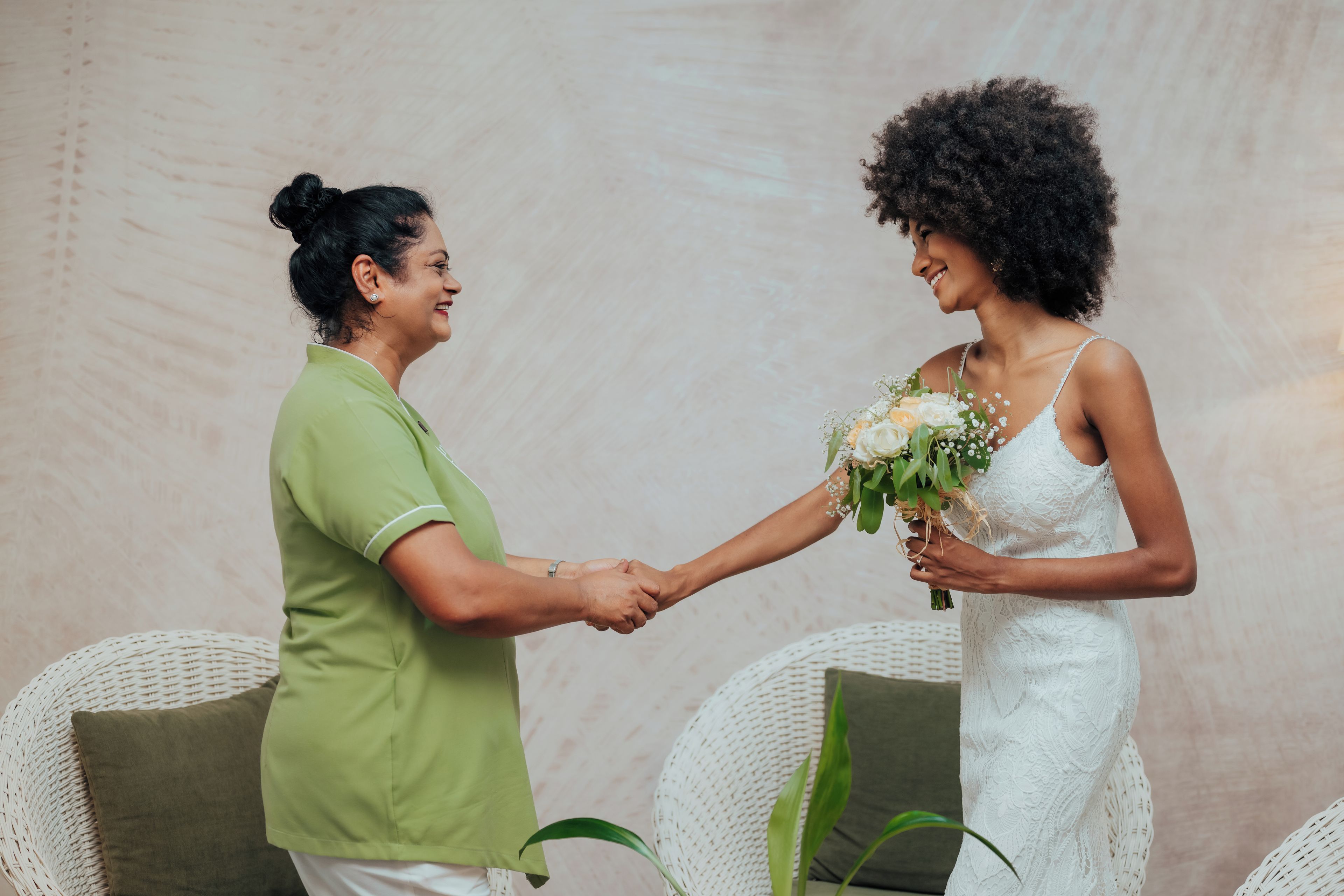 Two individuals holding hands, one in a green shirt and the other in a white dress, with wicker chairs and a plain wall in the background.
