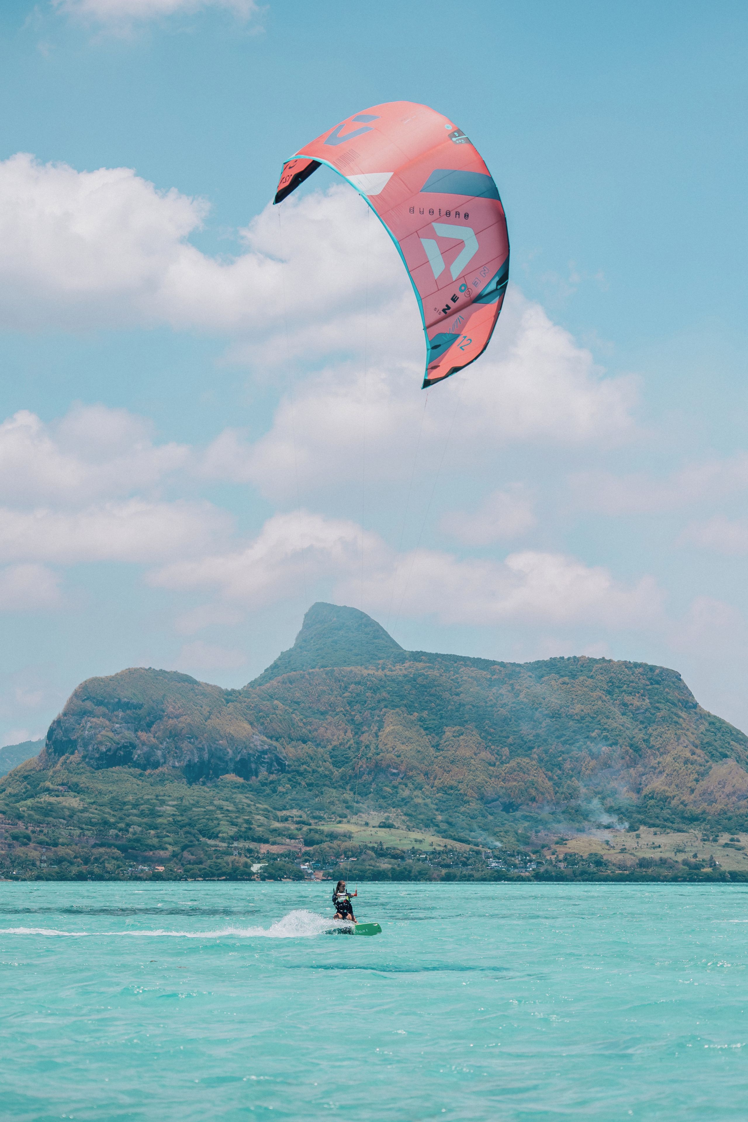 Una persona practica windsurf sobre aguas turquesas, con un paisaje montañoso al fondo.