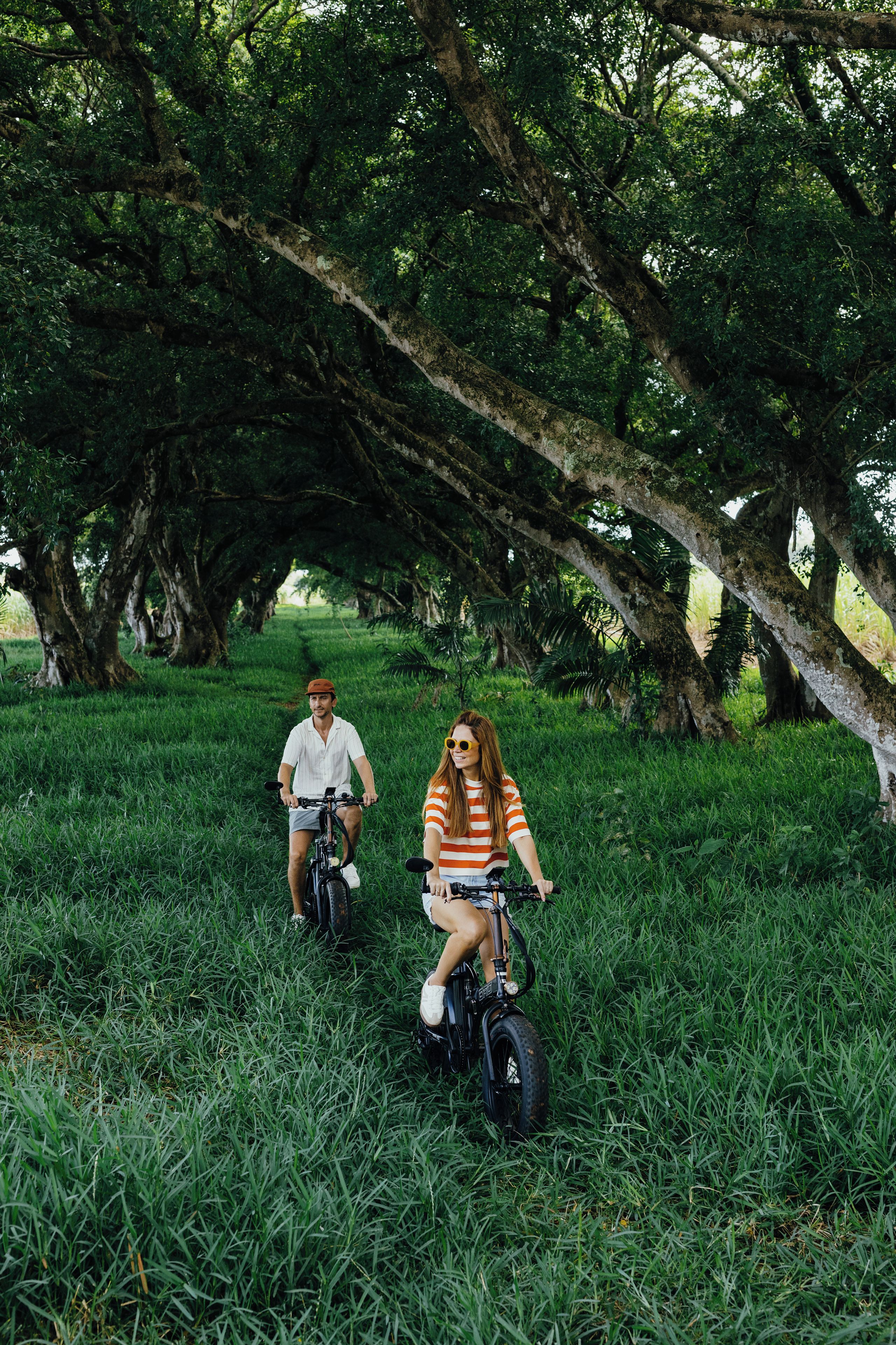 Two people riding electric bikes on a grassy path under large trees.