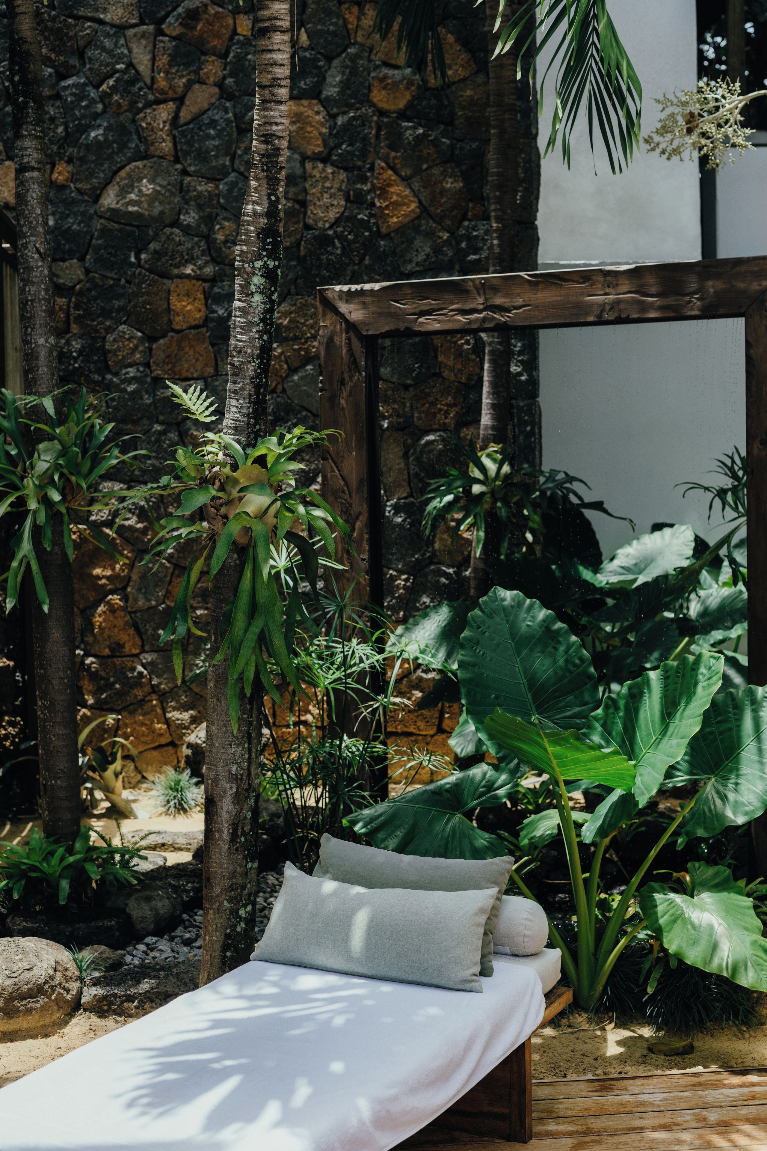 A bed with pillows in a room with a stone wall and tropical plants.