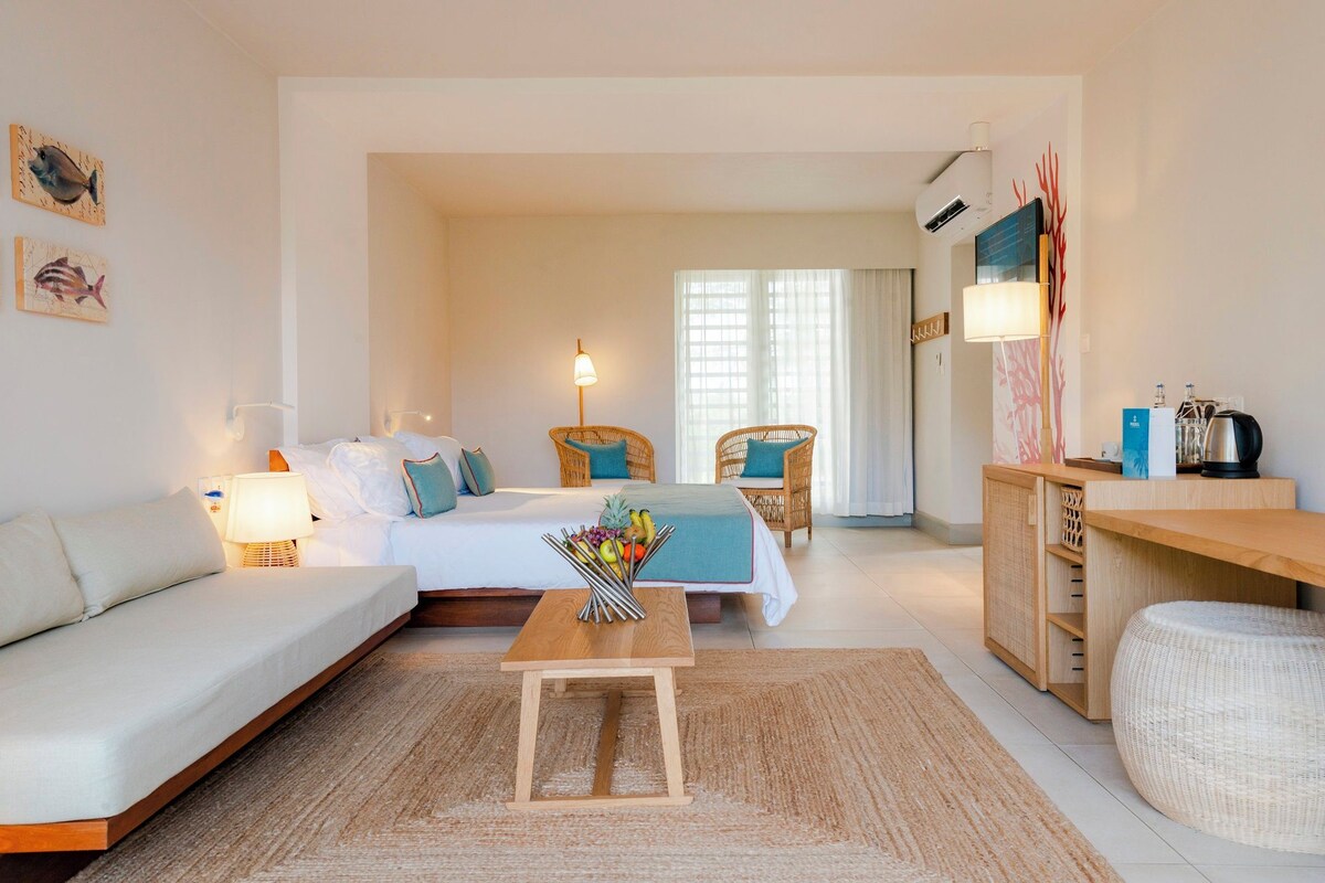 A modern hotel room with a bed, a sofa, a small table with fruit, wicker chairs, a desk with a kettle, and artwork on the walls.