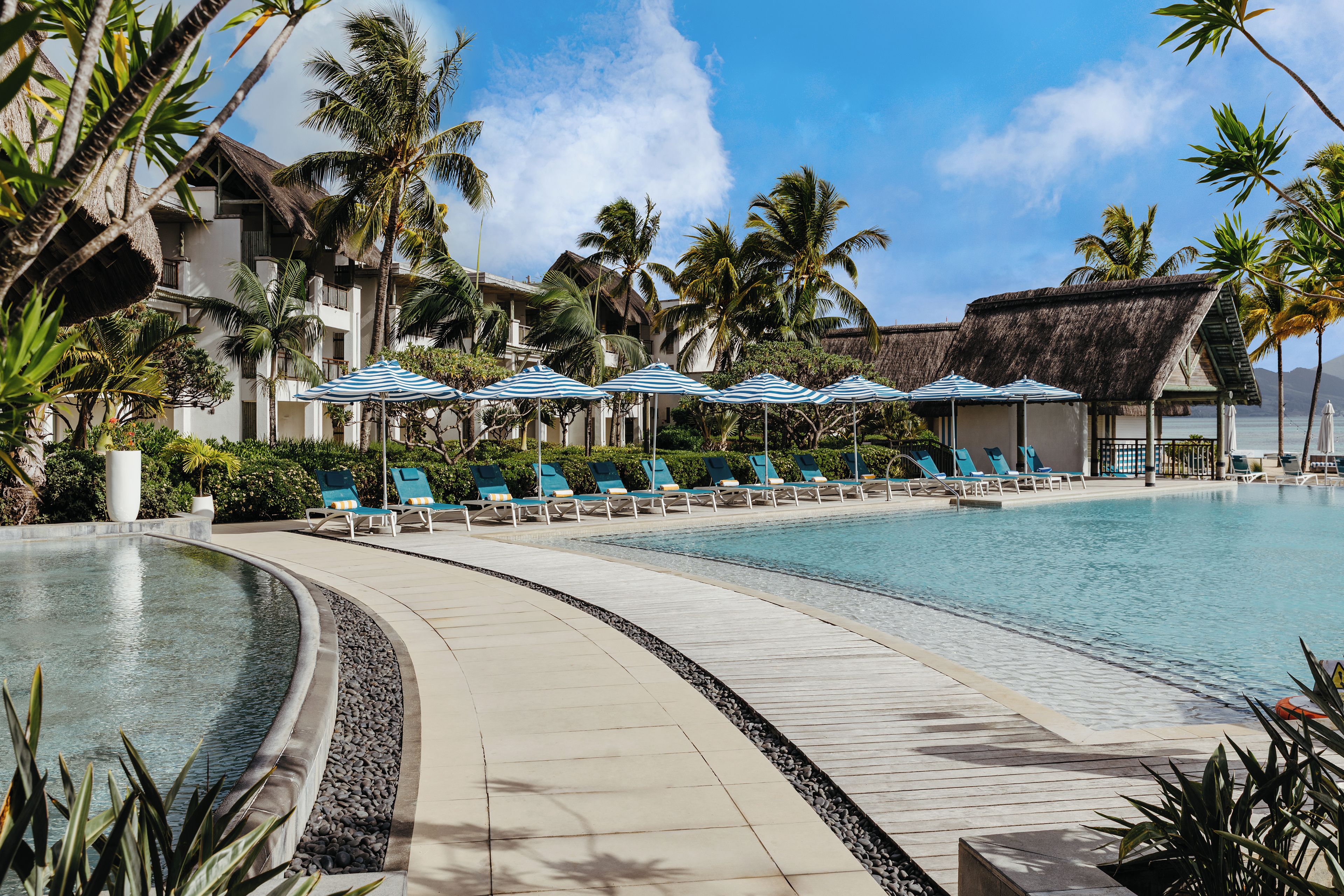 A resort pool area with lounge chairs and umbrellas, surrounded by palm trees and a building with a thatched roof.