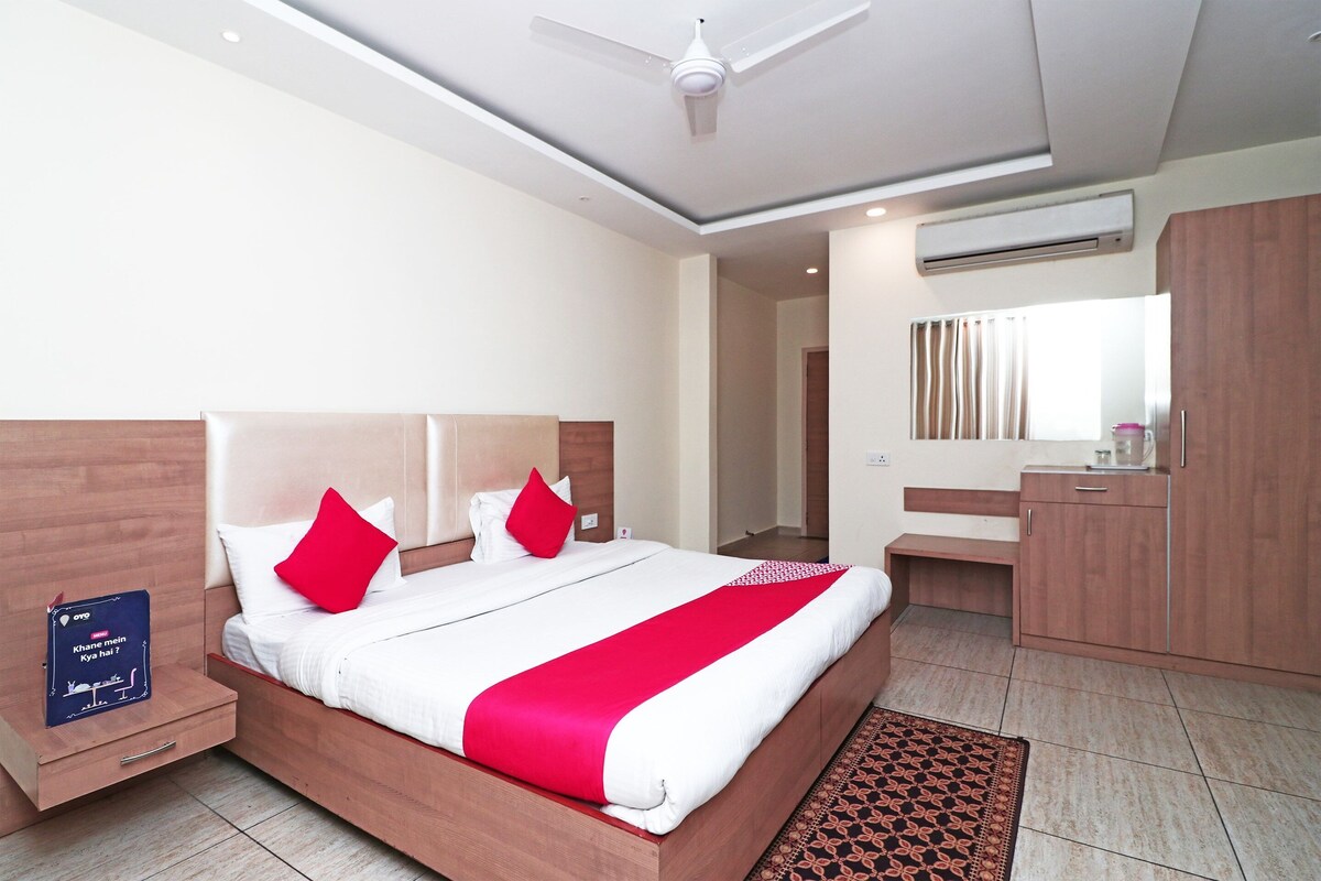 A modern hotel room with a large bed, wooden headboard, a bedside table, a nightstand, a red and white bedspread, a patterned rug, a ceiling fan, an air conditioning unit, a mirror, a wooden bench, and a small cabinet.