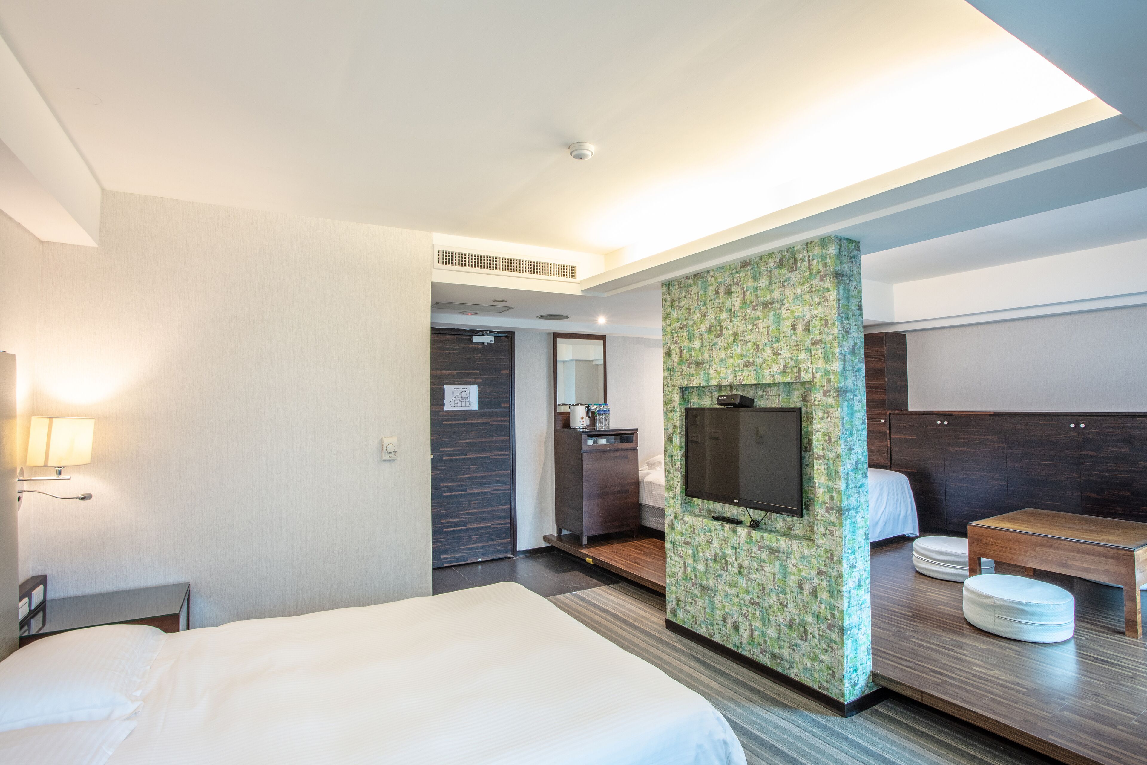 Une chambre d’hôtel comprenant un lit, un bureau, un téléviseur fixé au mur, qui est décoré d’un motif vert, une table de chevet en bois et une petite table avec un banc.