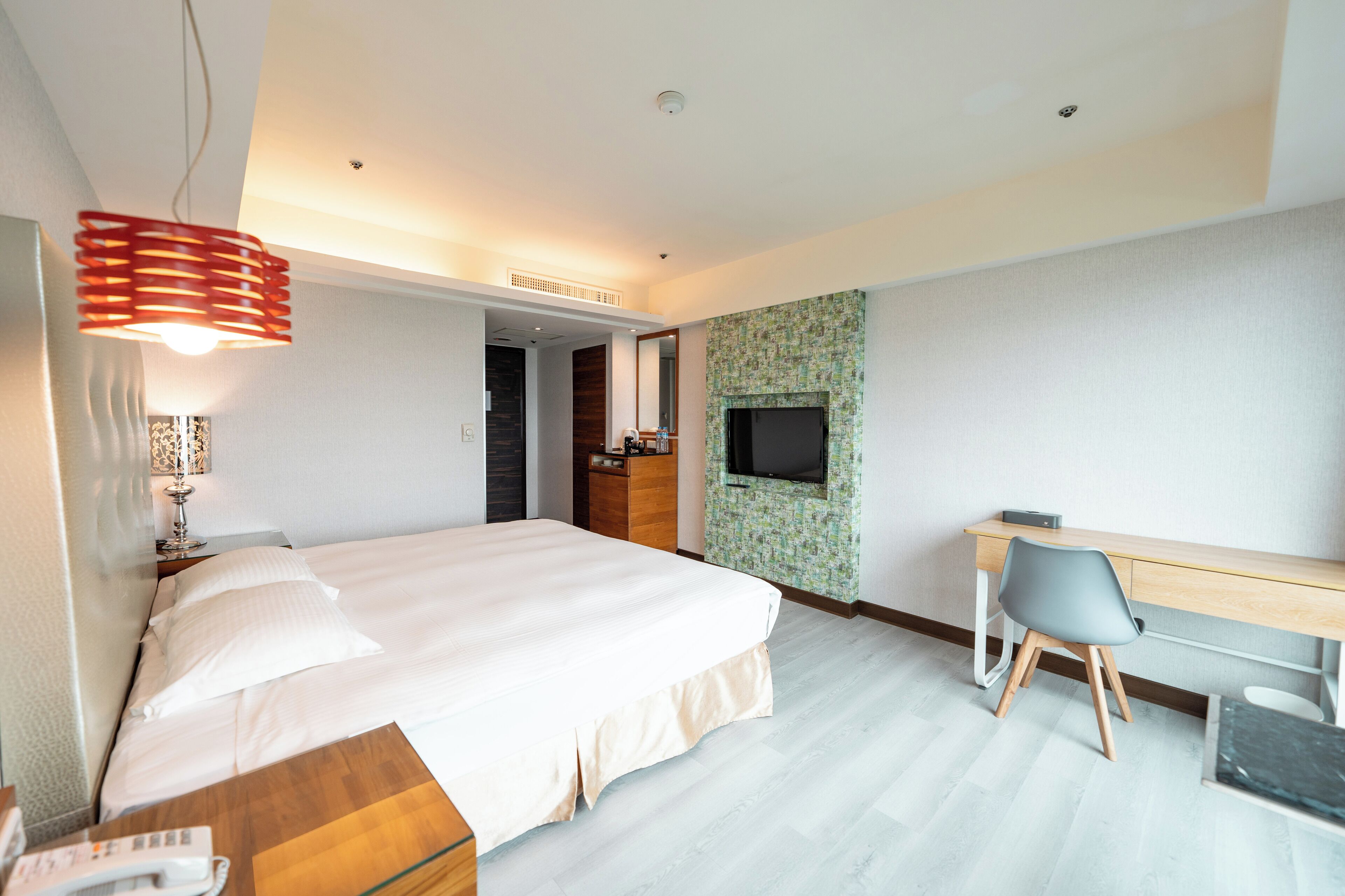 A modern hotel room with a large bed, a desk with a chair, a TV mounted on the wall, and a unique red pendant light.