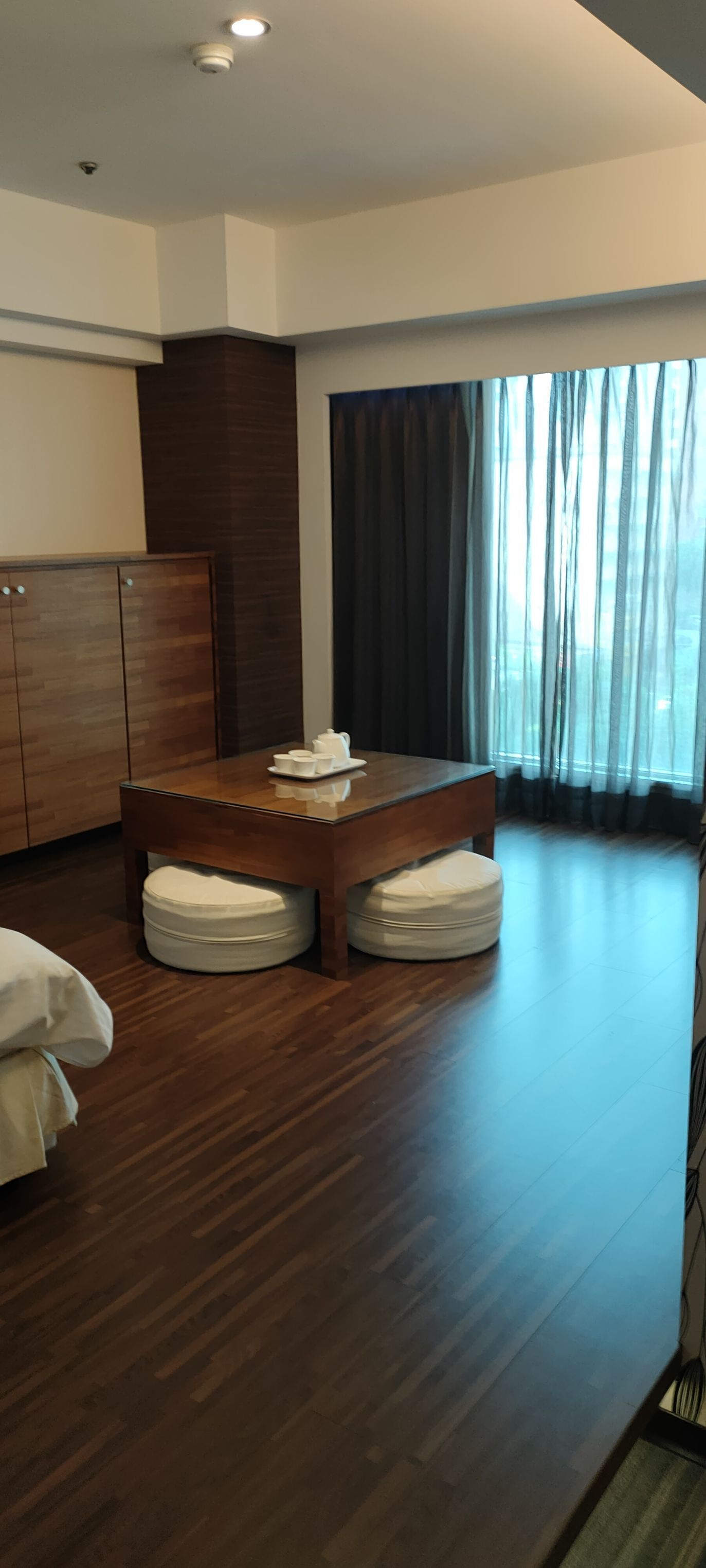 Quarto de hotel moderno com piso de madeira, uma janela grande com cortinas, uma mesa de madeira com um conjunto de chá e um guarda-roupa.