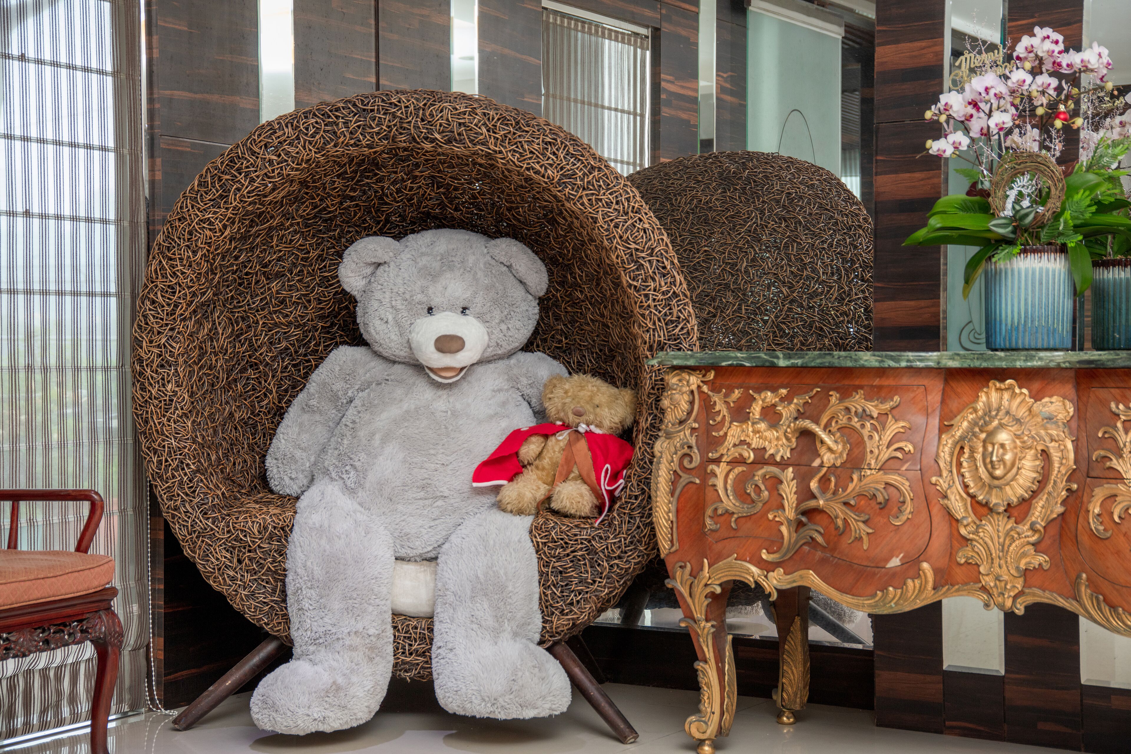 A large grey teddy bear seated in a wicker chair with a smaller brown teddy bear beside it.