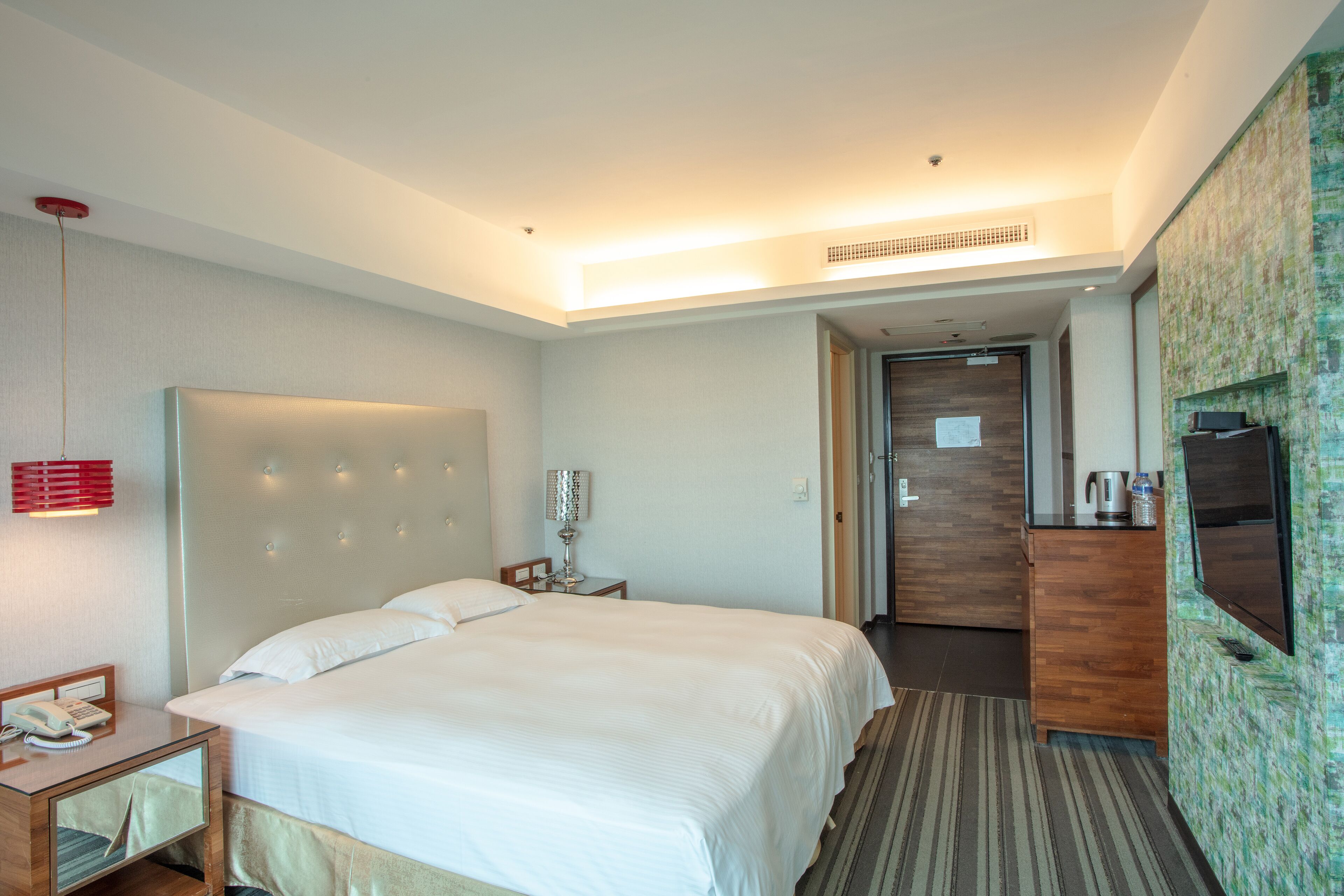 A hotel room with a large bed, bedside table with a lamp, a TV mounted on the wall, and a wooden dresser.