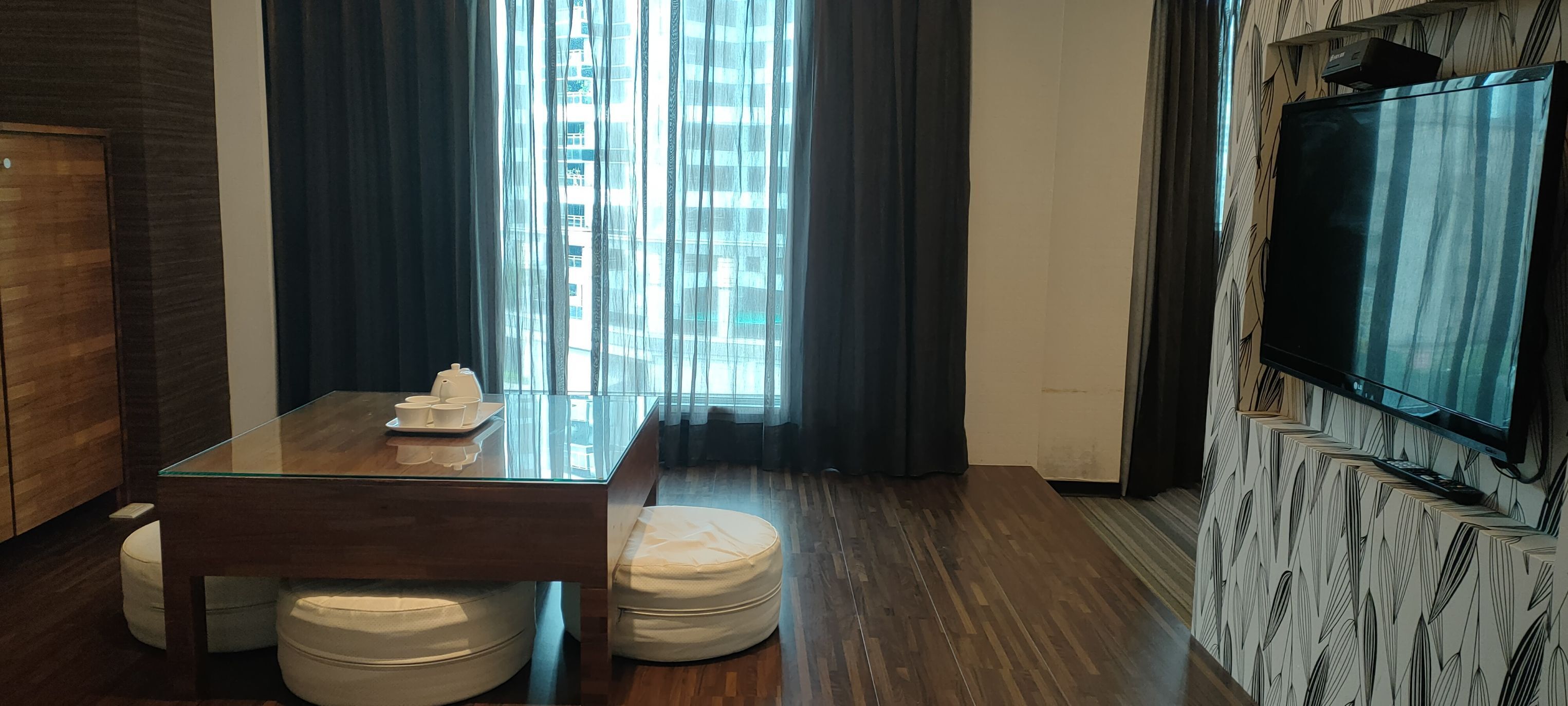 A modern hotel room with a glass dining table, two white ottomans, a flat-screen TV mounted on a patterned wall, and a large window with curtains.