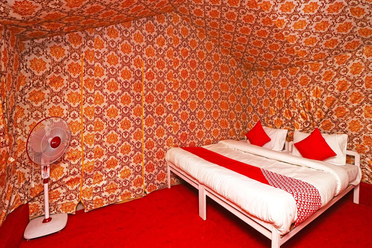 Une chambre avec un papier peint à motifs, un lit avec des coussins rouges et un ventilateur sur pied.
