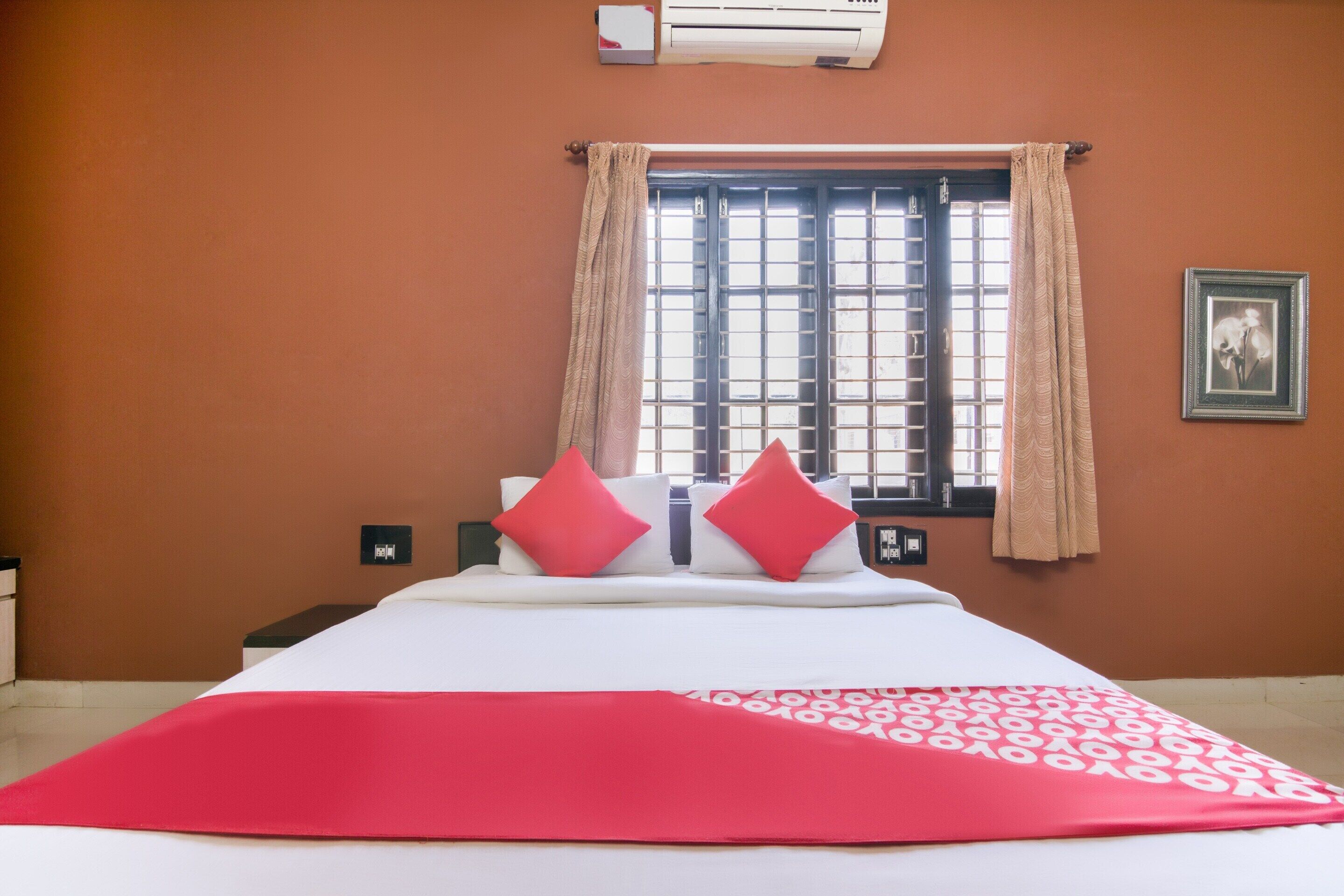 A hotel room with a bed, a window with curtains, an air conditioning unit, and a framed picture on the wall.