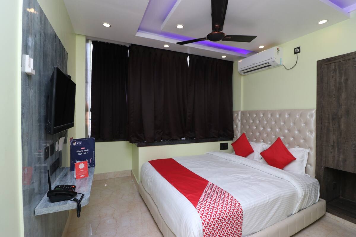 A hotel room with a bed, a TV, a desk with a phone, and an air conditioning unit.