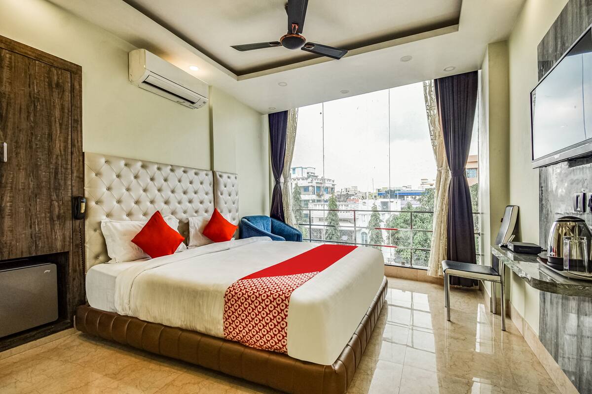A modern hotel room with a large bed, a ceiling fan, an air conditioning unit, a TV, and a view of the city through large windows.