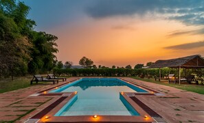 A rectangular pool with a wooden deck surrounding it, benches, and a thatched-roof structure. - Tiger Machan (Sawai Madhopur)