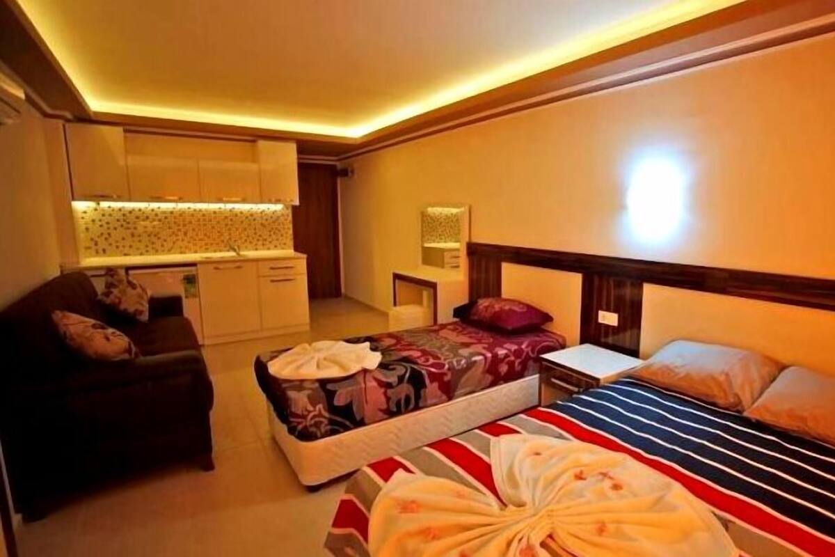 A hotel room with a bed, a sofa, a small table, a nightstand, a mirror, and a kitchenette area.