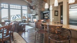 A spacious living area with a stone fireplace, a bar with copper bar stools, and a large window offering a mountain view. - Moonlight Basin | Ski-In/ Out | Hot Tub (Big Sky)
