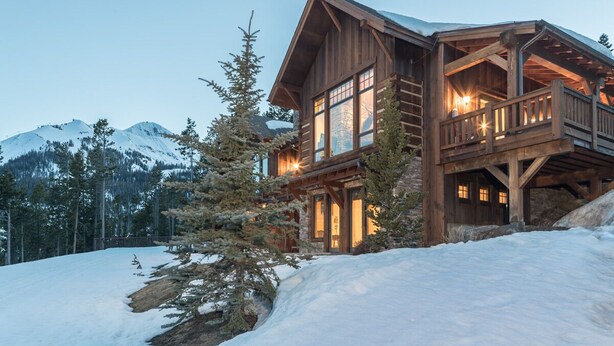 A wooden cabin with a balcony, surrounded by snow and trees, with a mountain in the background. - Moonlight Basin | Ski-In/ Out | Hot Tub (Big Sky)