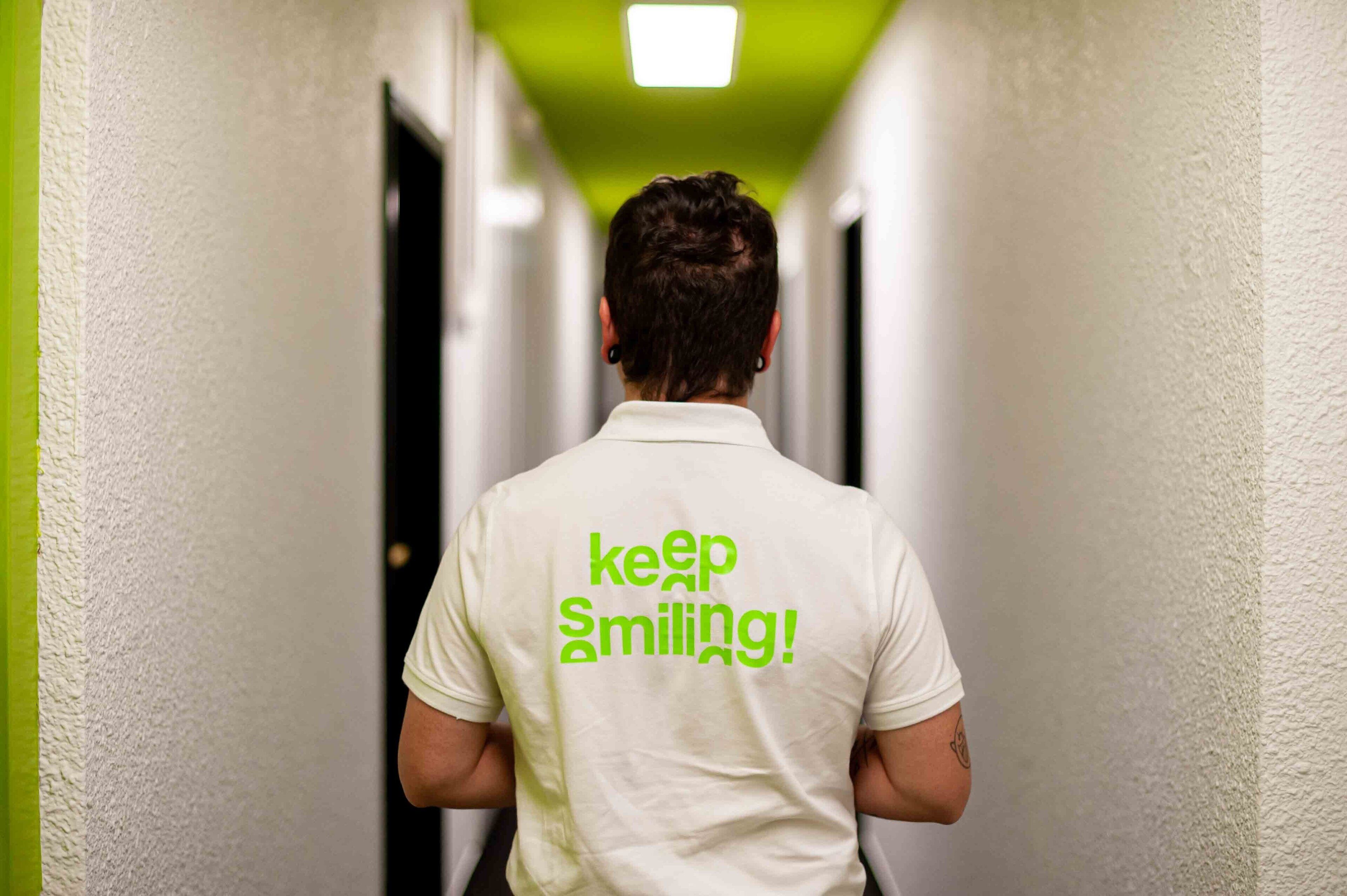 Eine Person geht einen Flur entlang, auf deren Hemd ein Schild mit der Aufschrift 'Keep Smiling!' befestigt ist.
