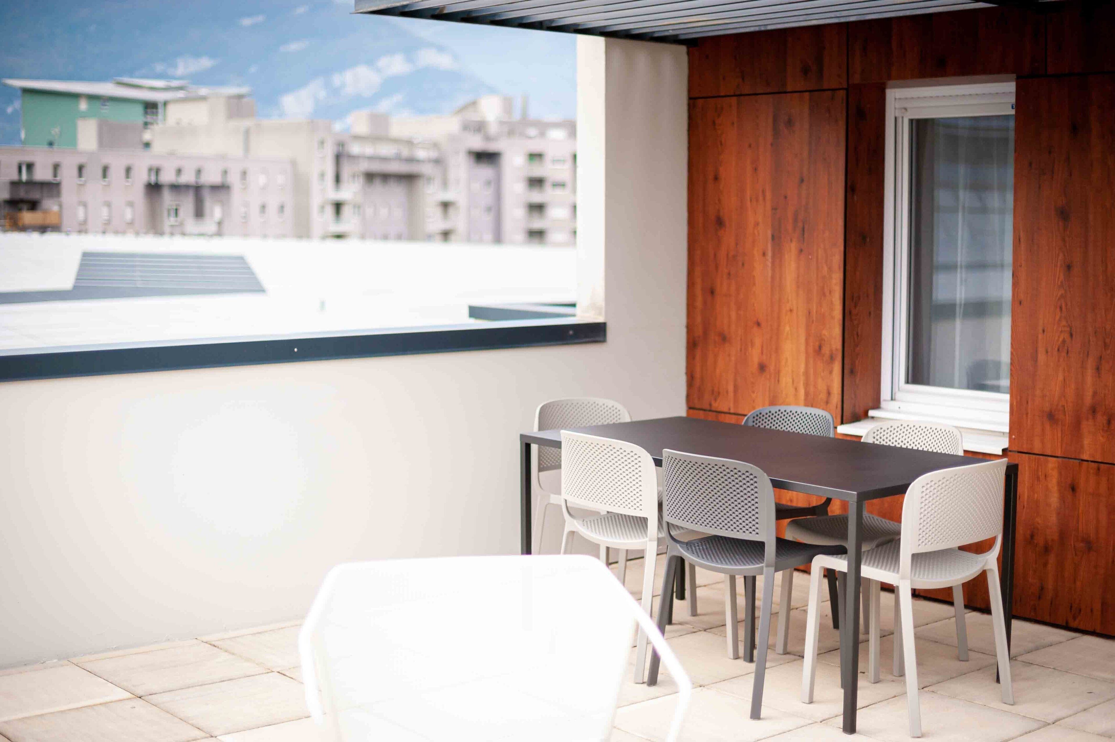 A balcony with a table and chairs, a view of buildings, and a wooden wall.