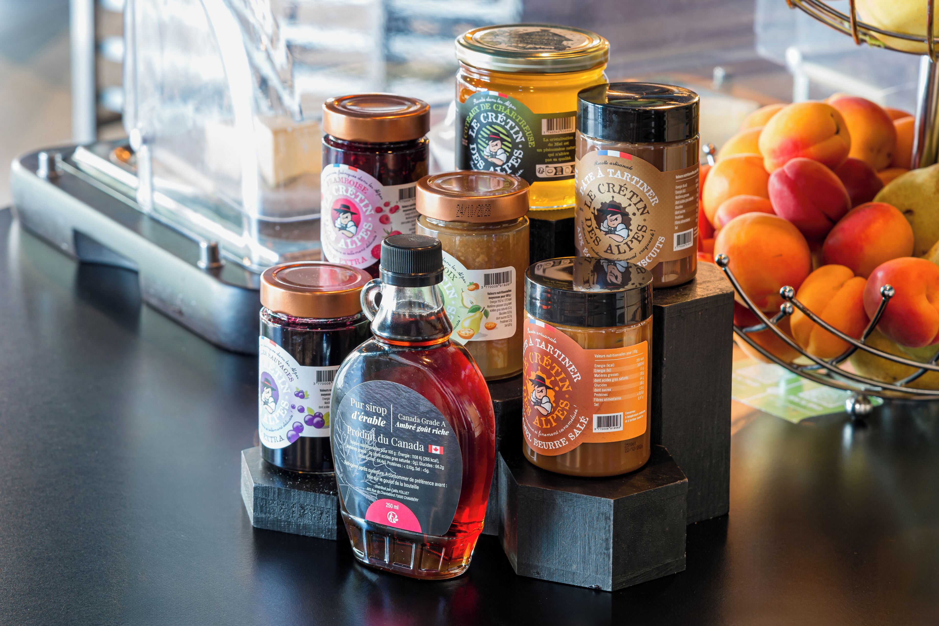 Une sélection de produits alimentaires, notamment des pots de confitures et une bouteille de sirop, est exposée sur un comptoir, avec une variété de fruits en arrière-plan.
