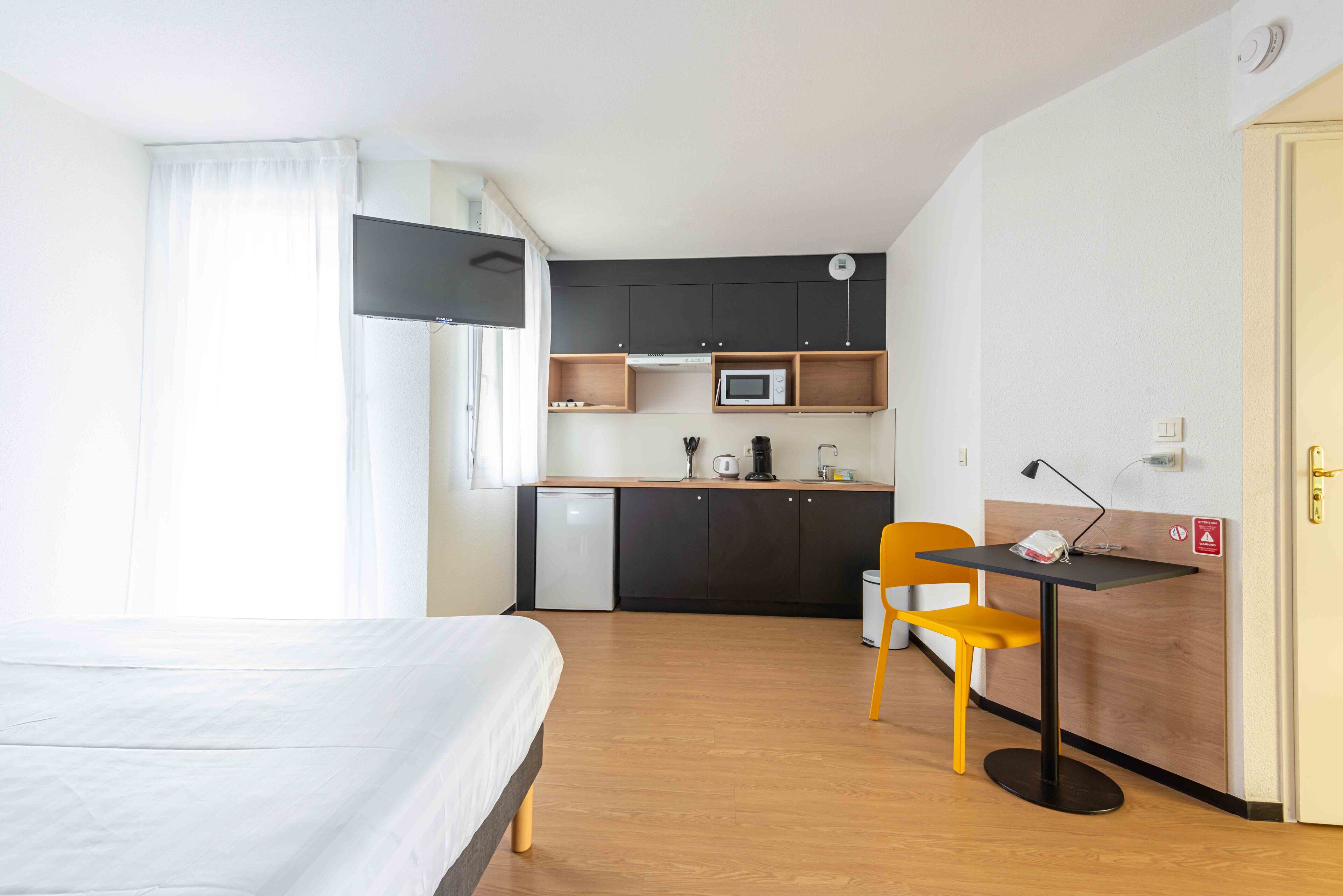 Ein kleines Hotelzimmer mit Bett, einer kompakten Küchenzeile, einem Schreibtisch mit Stuhl und einem an der Wand befestigten Fernseher.