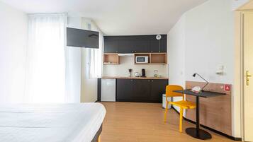 Ein kleines Hotelzimmer mit Bett, einer kompakten Küchenzeile, einem Schreibtisch mit Stuhl und einem an der Wand befestigten Fernseher.