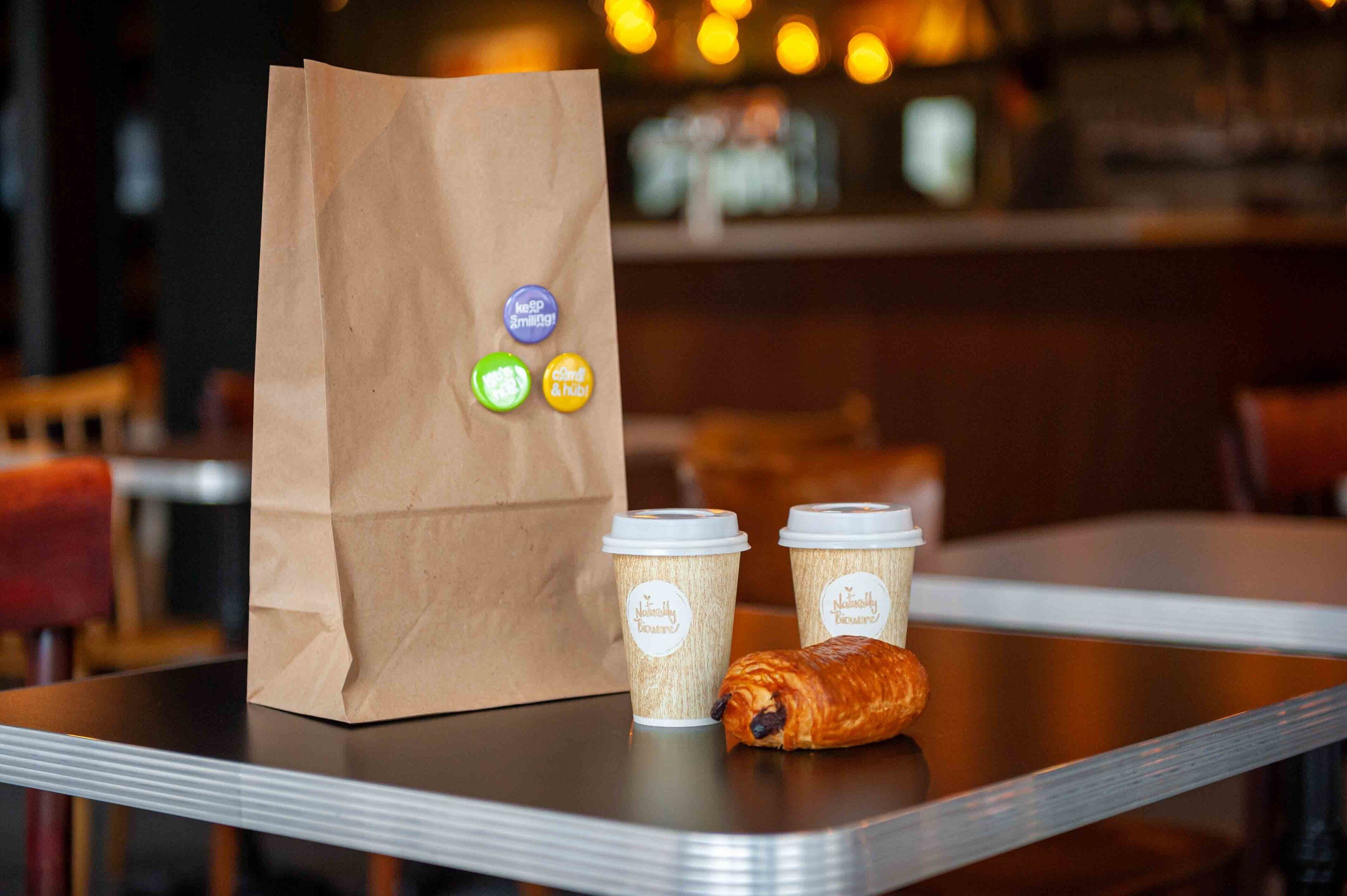 Eine Papiertüte mit Recyclingzeichen, zwei Kaffeetassen und ein Croissant auf einem Tisch.