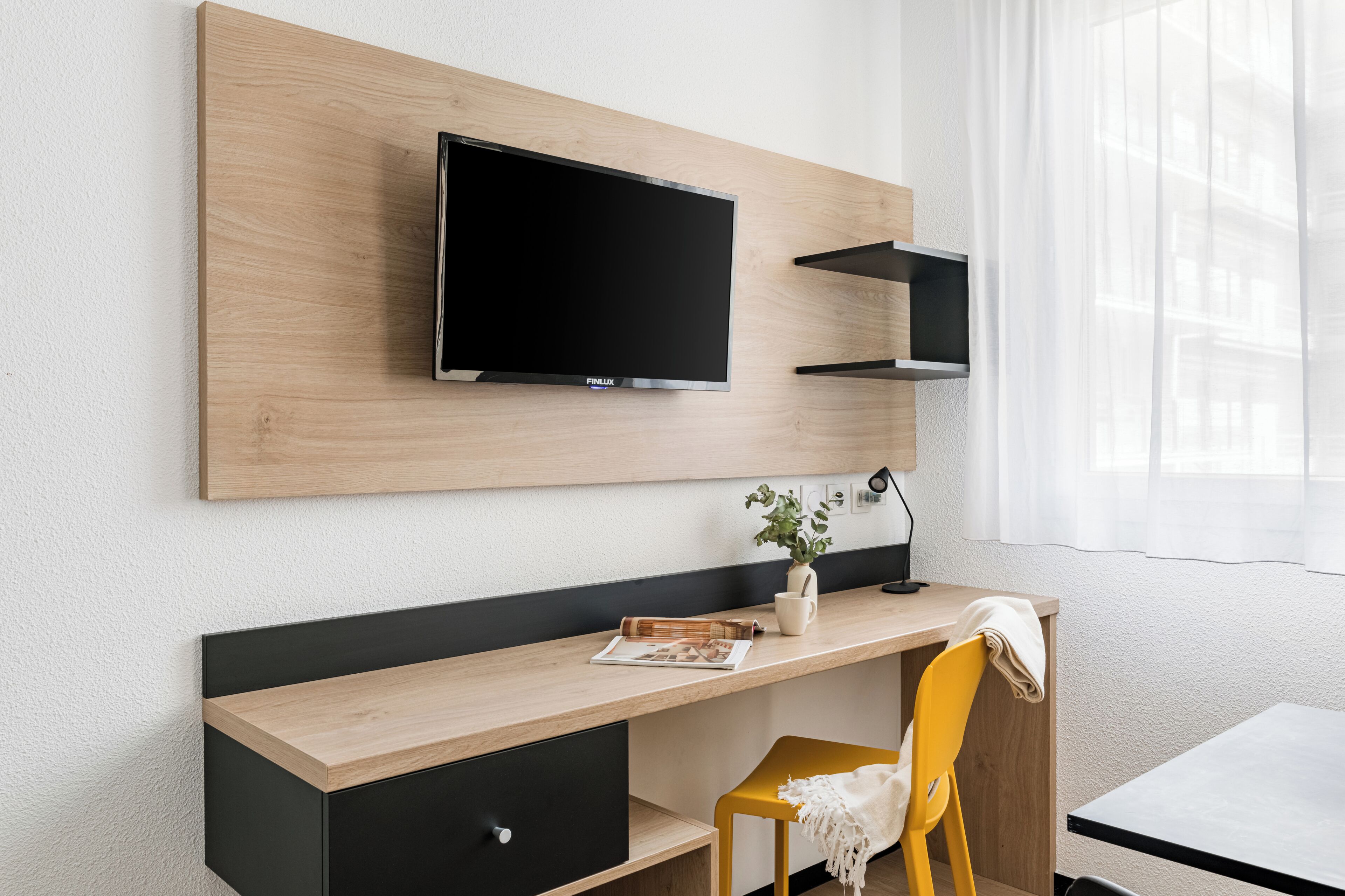 Ein modernes Hotelzimmer mit Schreibtisch aus Holz, einem Flachbildfernseher, einer kleinen Pflanze und einem gelben Stuhl.