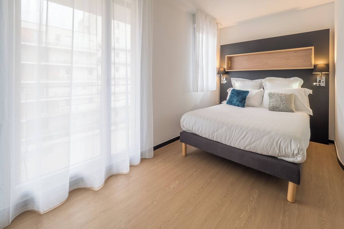 Een moderne slaapkamer met een groot bed, een houten hoofdbord en een raam met dunne gordijnen.