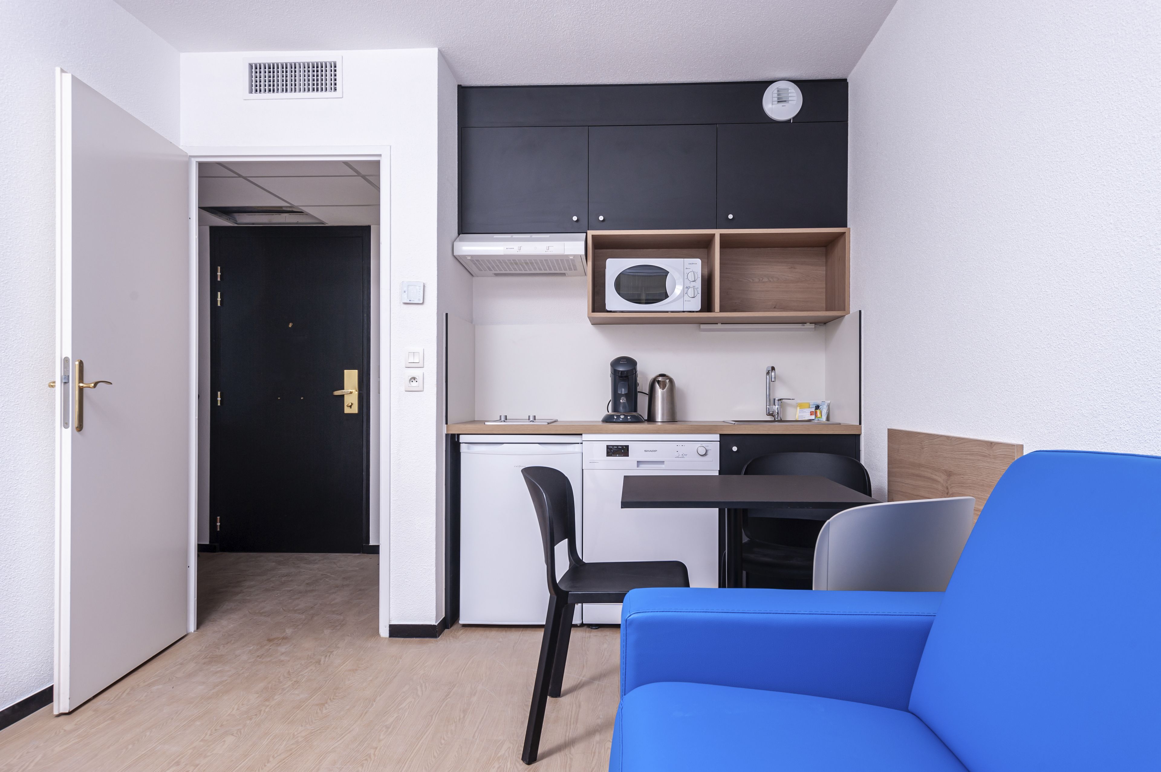 Een compacte kitchenette met een magnetron, koffiezetapparaat en vaatwasser. In de woonruimte staat een blauwe bank en een zwarte fauteuil.