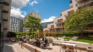 Un espacio al aire libre con patio, mobiliario y mesas, rodeado de vegetación y edificios modernos.