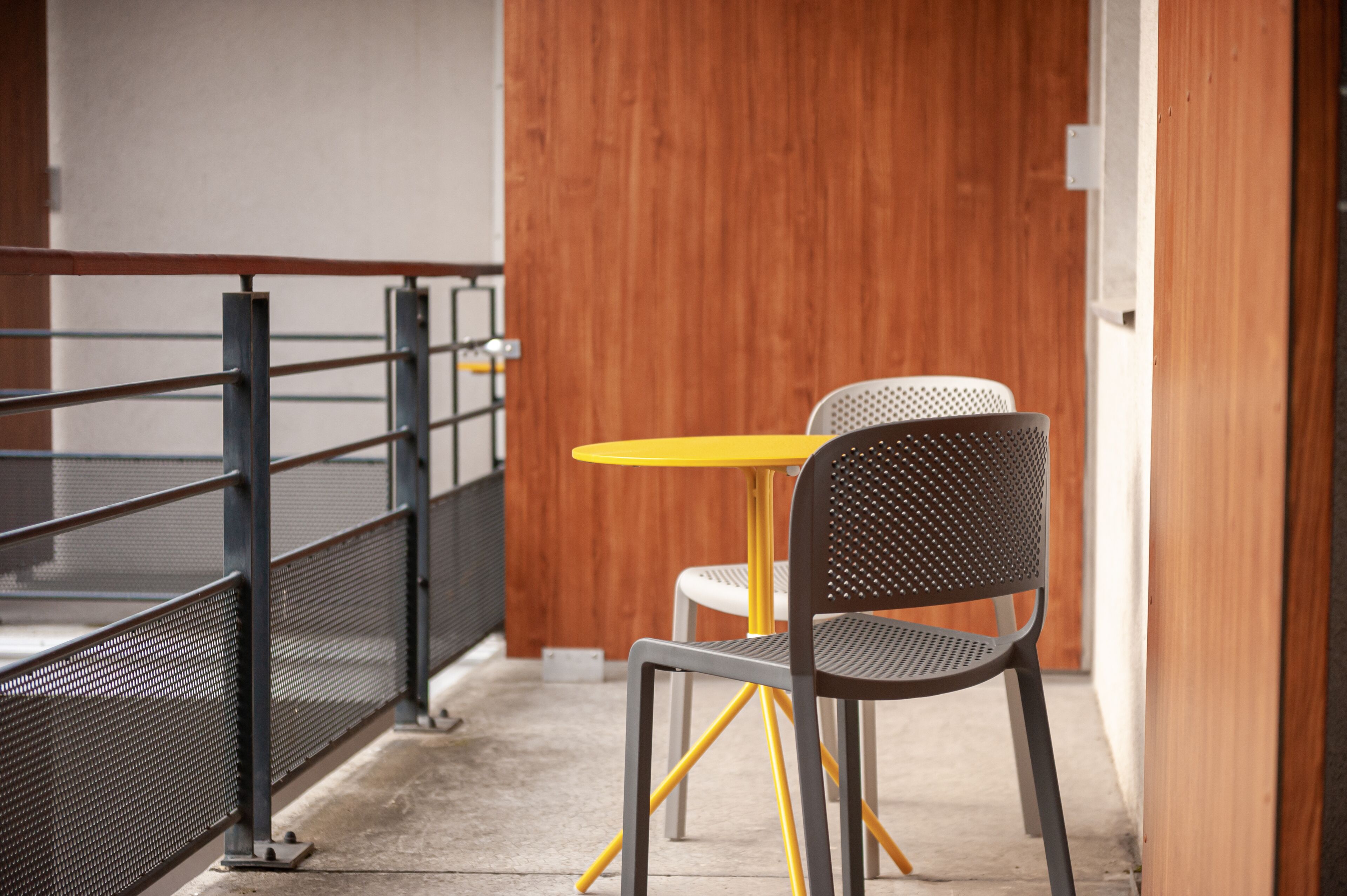 A small outdoor seating area with a metal railing, a yellow table, and two chairs.