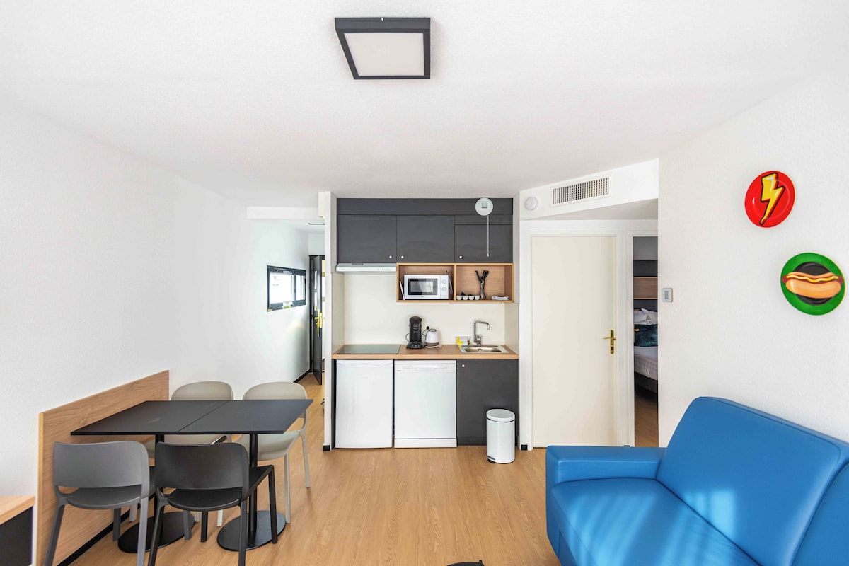 Un espacio habitable compacto con kitchenette, una zona de comedor con mesa y sillas, y un sofá azul.