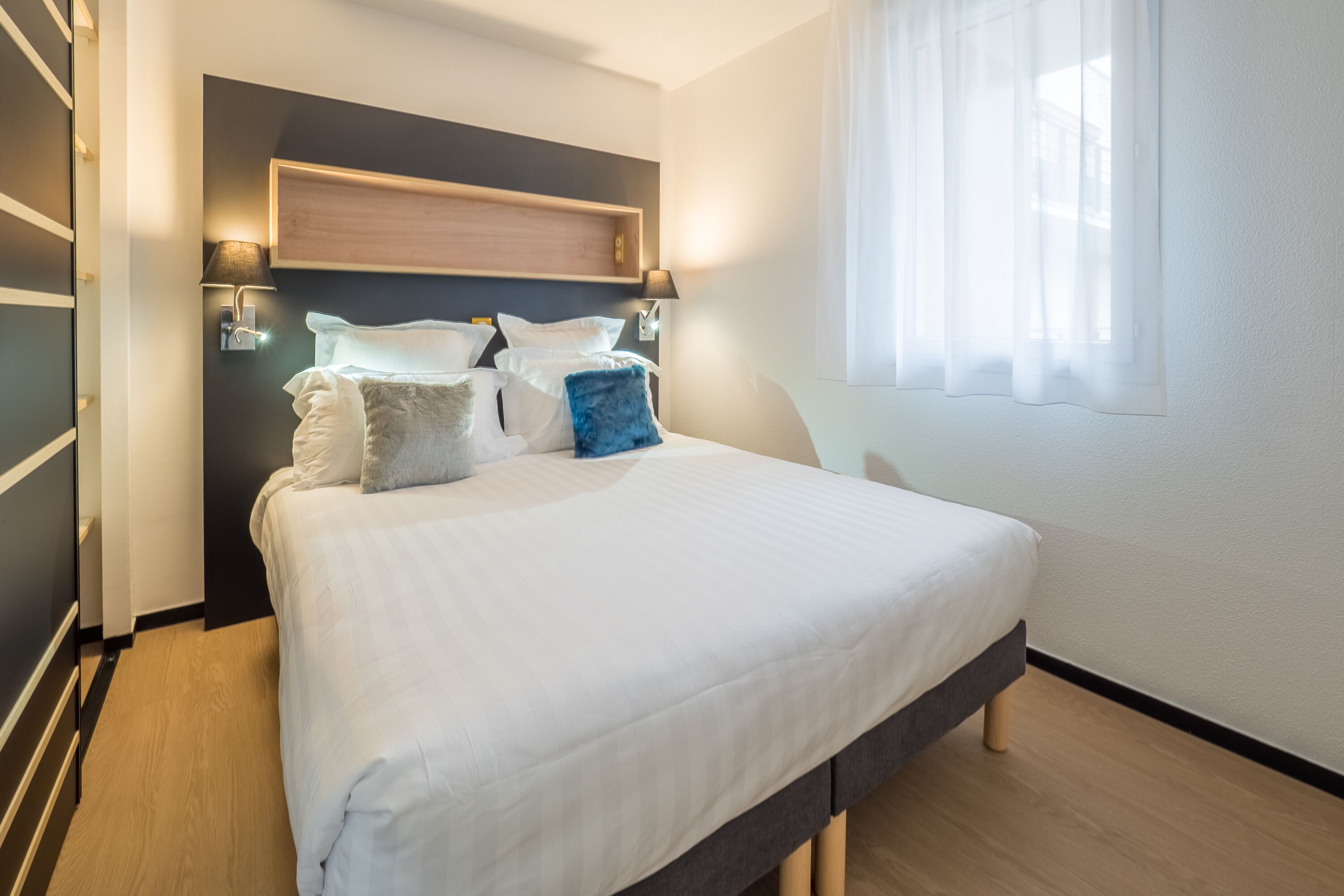 Ein modernes Schlafzimmer mit einem großen Bett, zwei Wandlampen und einem Fenster mit durchsichtigen Vorhängen.