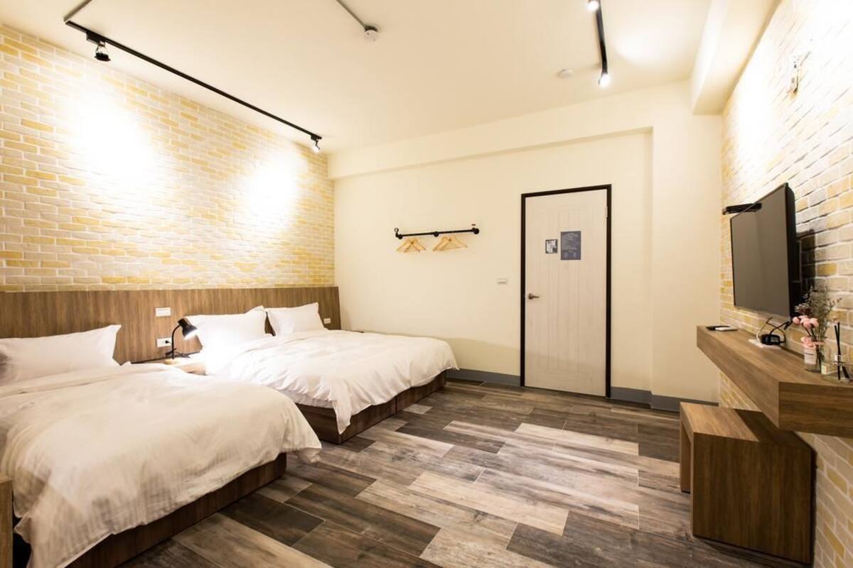 A hotel room with two beds, a wooden headboard, a brick wall, a wooden floor, a TV, and a wooden counter with a mirror.