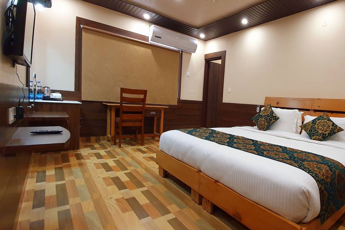 A hotel room with a wooden bed, a desk with a chair, a bedside table, and a large window.