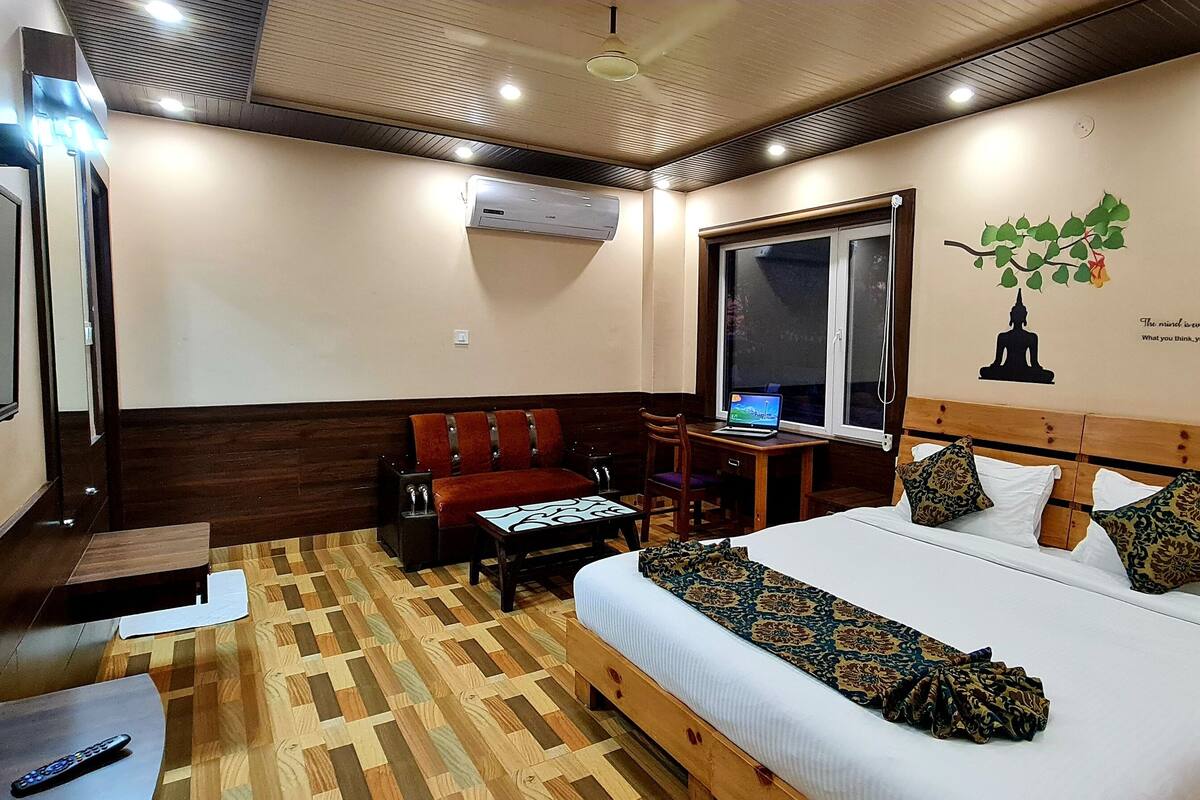 A hotel room with a bed, a desk with a computer, a chair, a TV, and an air conditioning unit.