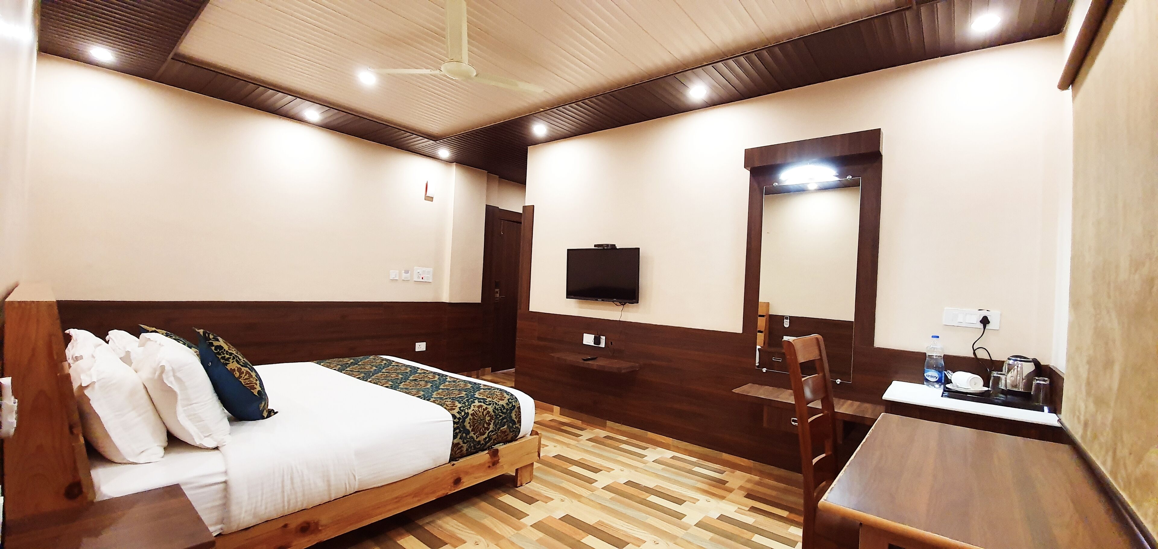 A hotel room with a wooden bed, a desk with a chair, a mirror, and a television on the wall.