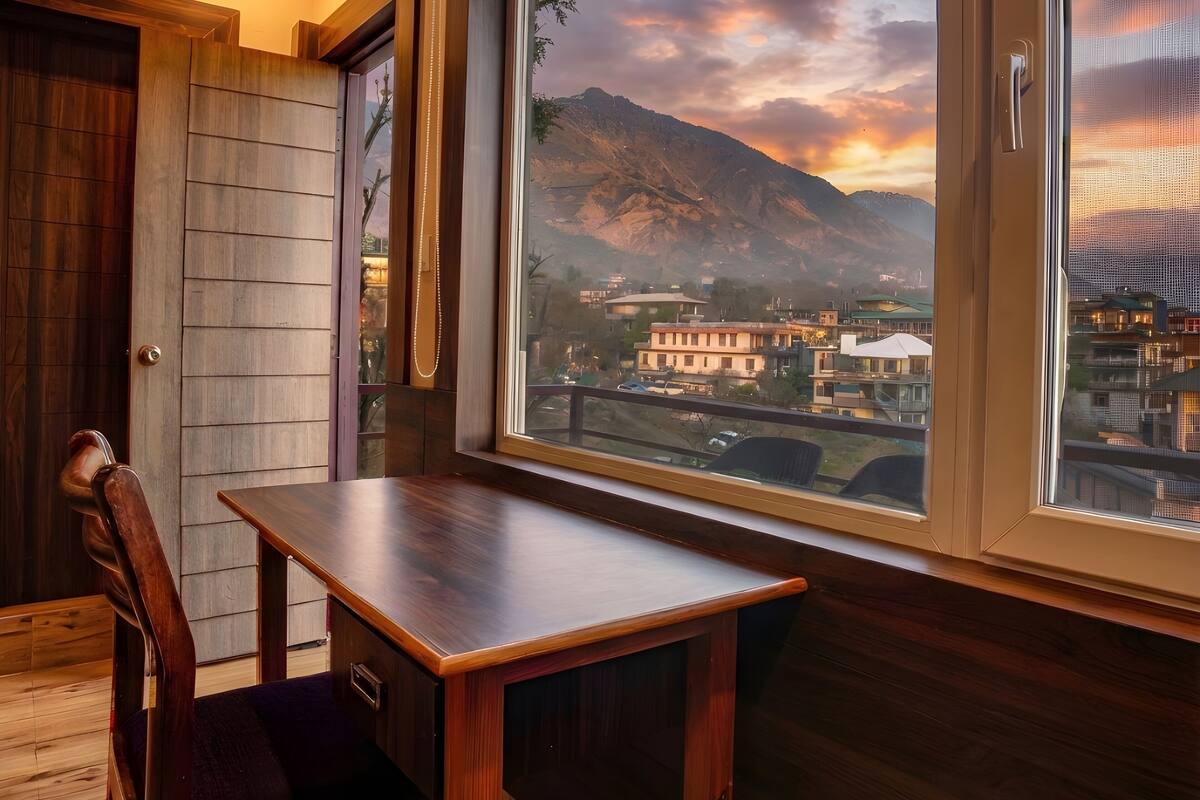 A corner of a room with a wooden table and chair, a large window with a view of a mountain and buildings, and a door with a wooden panel.