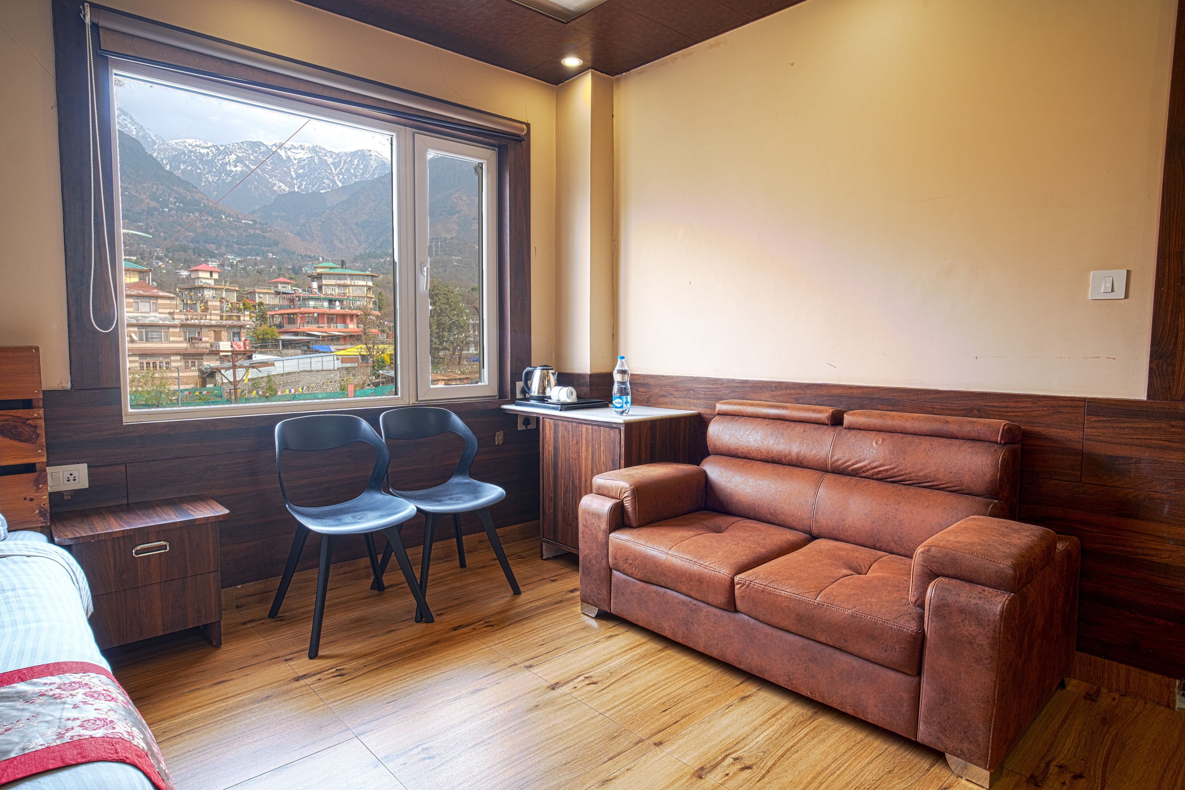 A room with a wooden floor, a brown leather sofa, two chairs, a small table with a kettle, and a window with a mountain view.