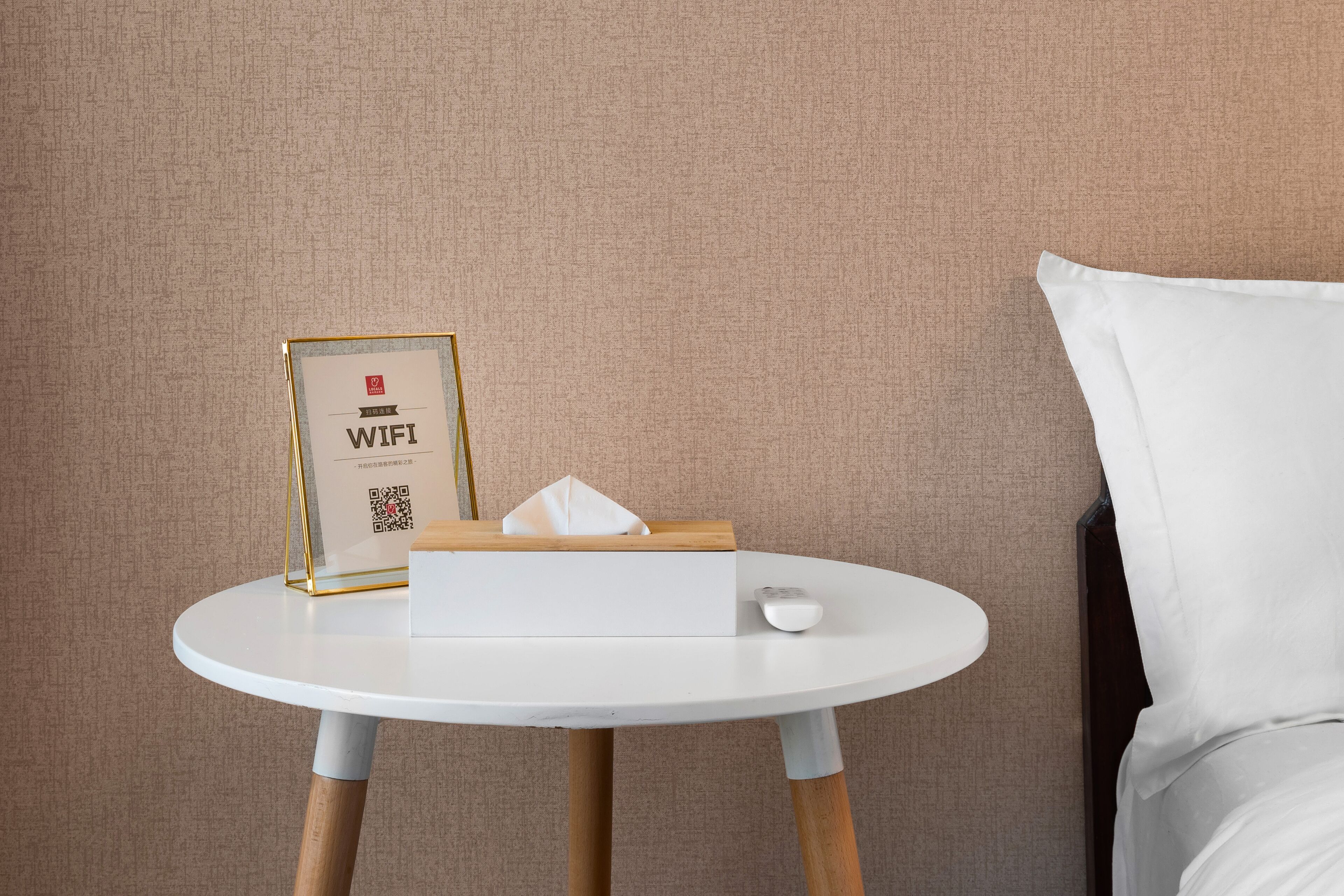 Ein ordentlich eingerichtetes Hotelzimmer mit einem runden weißen Tisch, der ein gerahmtes Schild mit der Aufschrift 'WiFi', eine Taschentuchbox und eine Fernbedienung zeigt.