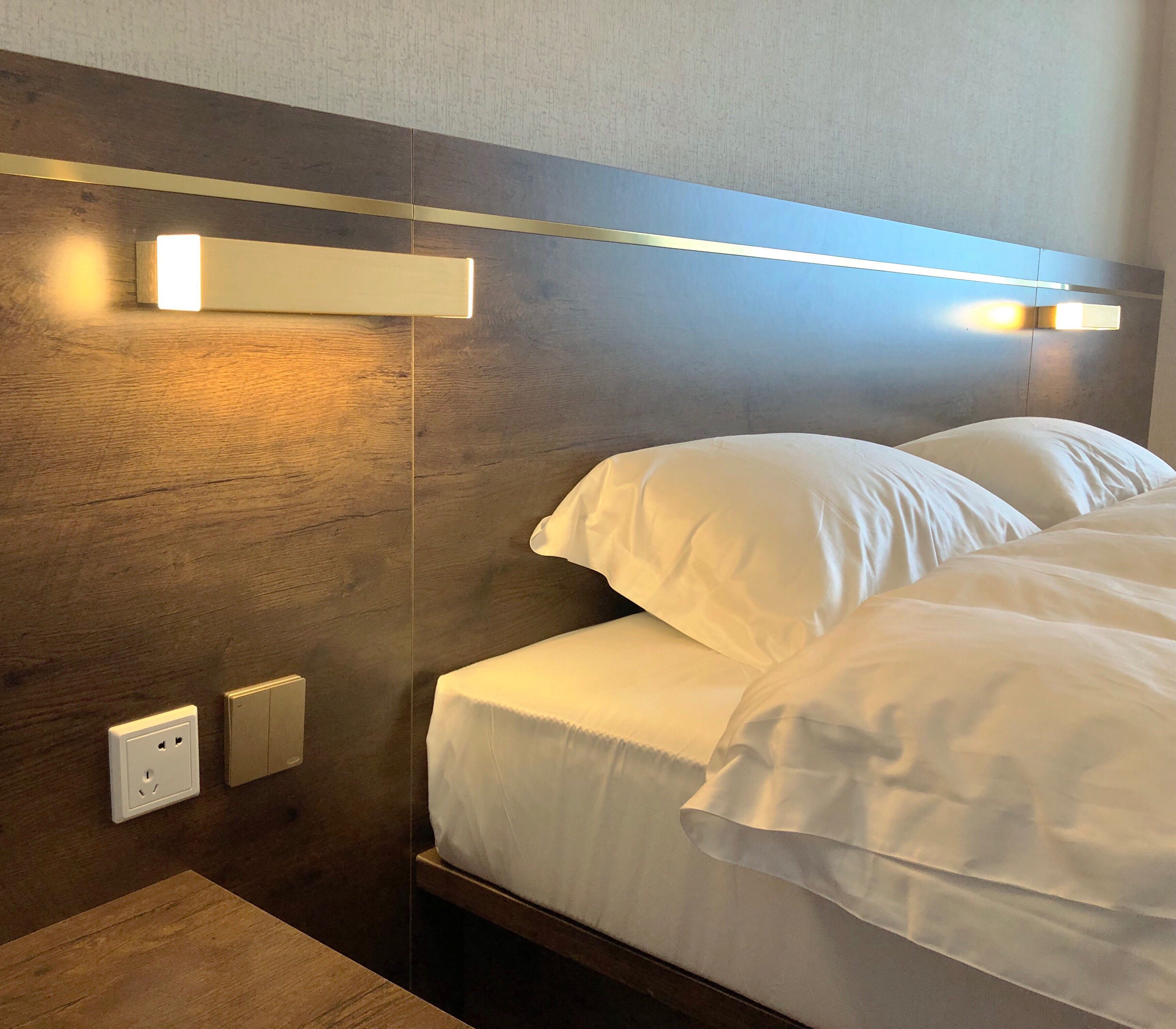 Een hotelkamer met een houten hoofdeinde, twee witte kussens en een nachtkastje met een lamp.